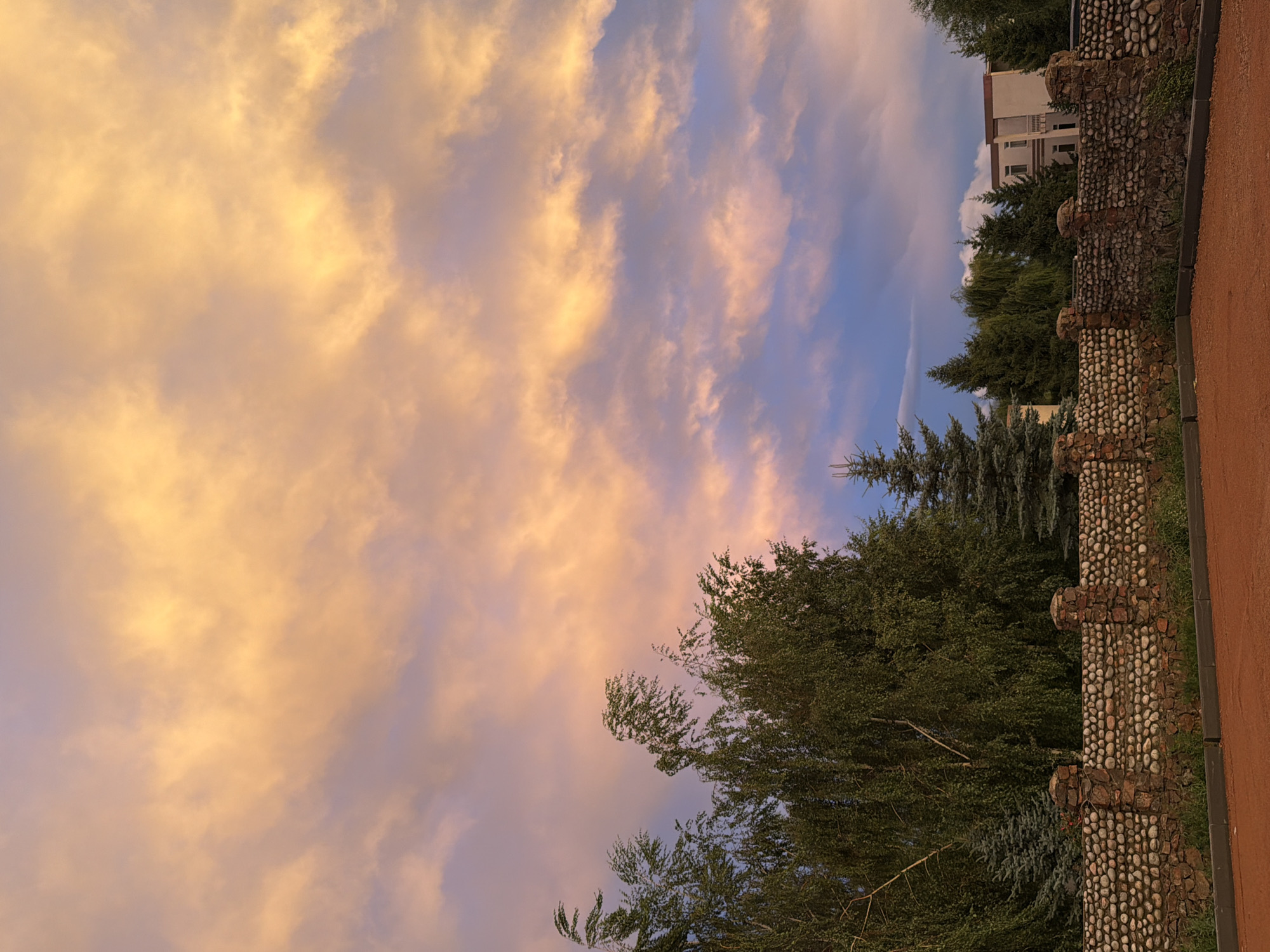 A low-angle, wide shot captures a segment of a rustic stone wall in the foreground, topped with rough-hewn stone pillars. Beyond the wall, a variety of trees, including tall, leafy deciduous trees and dark green conifers, are visible. In the background, a modern building with white and beige facades stands amongst the trees. The sky above is a canvas of dramatic clouds, predominantly illuminated in warm hues of orange and yellow, with hints of purple and blue peeking through. The image appears to be taken during sunset or sunrise, casting a soft, ambient light on the scene. The ground in front of the wall is a reddish-brown, appearing to be a dirt or gravel surface. There are no people or animals visible in the frame. The overall impression is one of a peaceful, natural setting contrasted with a modern structure, under a vibrant sky. The scene is likely located in or around the city of Cholpon-Ata, Kyrgyzstan, given the visual characteristics of the architecture and vegetation.