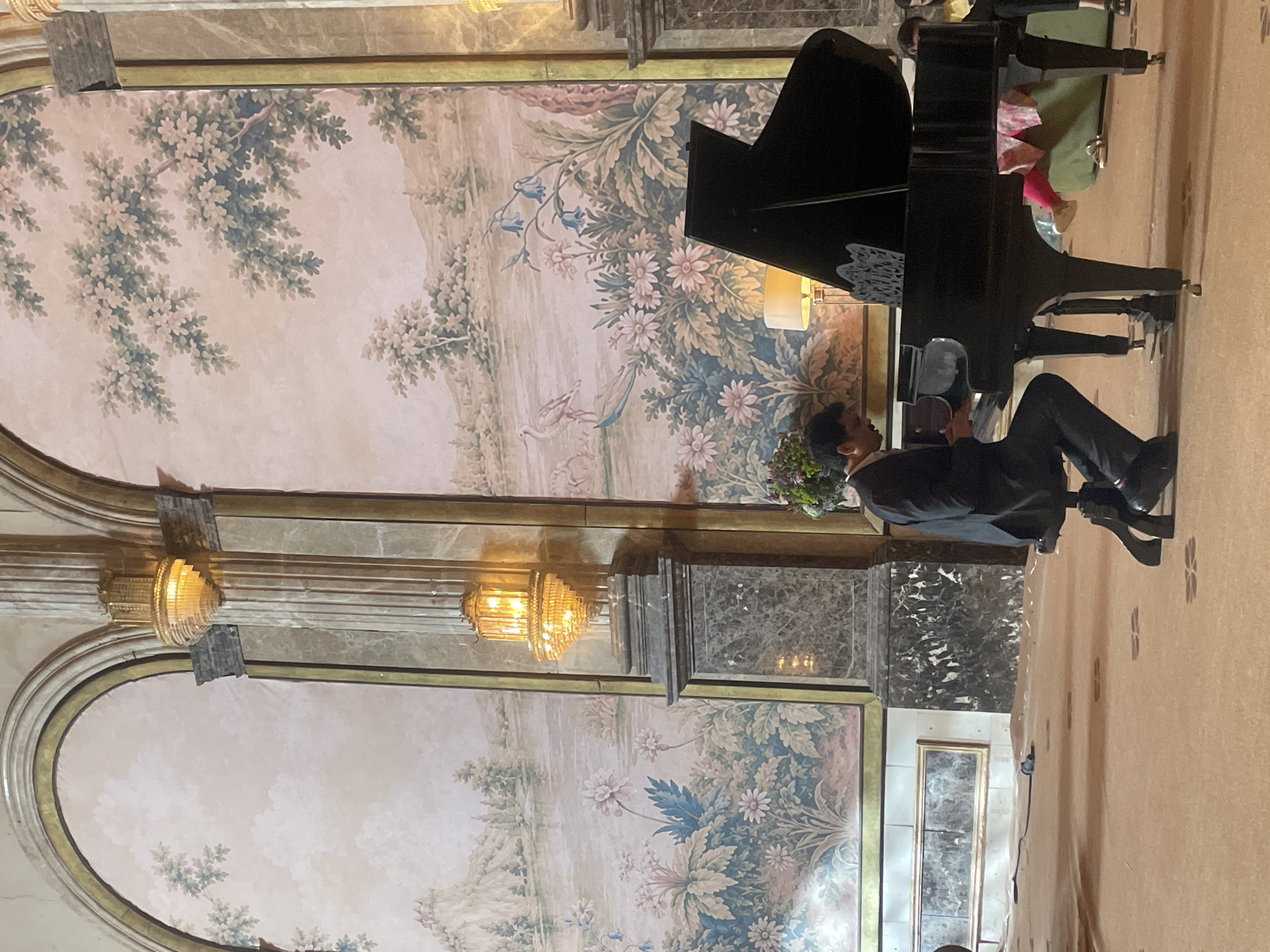 A young man, dressed in a dark suit and black shoes, sits on a bench in front of a grand piano, his hands poised over the keys. The piano, a glossy black instrument, is positioned in a room with ornate, classical decor. The walls are adorned with a large, faded mural depicting a pastoral landscape with trees, flowers, and birds. Two ornate chandeliers, with warm yellow lights, hang from the ceiling.
The lighting suggests it might be evening or an indoor setting with artificial light. The overall atmosphere appears formal and serene, possibly during a musical performance or practice session. The man's posture and focus suggest concentration.
The setting appears to be an indoor venue, possibly a concert hall, a ballroom, or a well-appointed room in a historic building. The carpeted floor and the detailed wall murals point to a luxurious environment. There is no visible text in the image. The context of Mumbai, India, is provided but not directly inferable from the visual cues in the image itself.