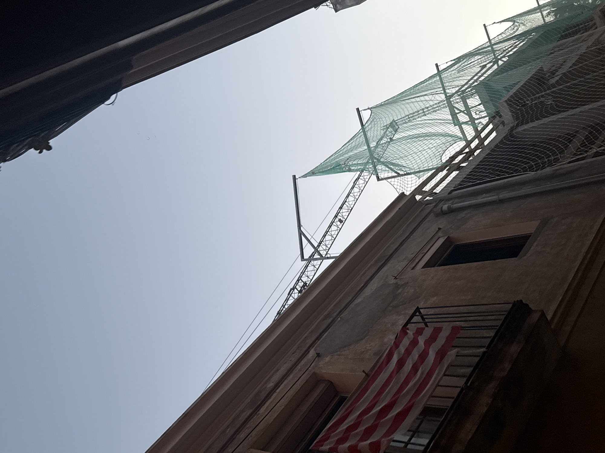 The image shows a close-up, low-angle shot of a building under construction or renovation. A balcony on the left side of the frame displays a flag with red and white horizontal stripes, possibly the flag of the Basque Country or a similar regional flag. Above the building, a construction crane is visible, along with green safety netting stretched across the scaffolding. The sky is clear and a pale blue, suggesting it is either early morning or late afternoon. The architecture of the building appears to be old, with weathered plaster. The scene is set in an urban environment, implied by the presence of buildings and construction. There are no people visible in the image. The location is identified as Sant Boi de Llobregat, Spain.