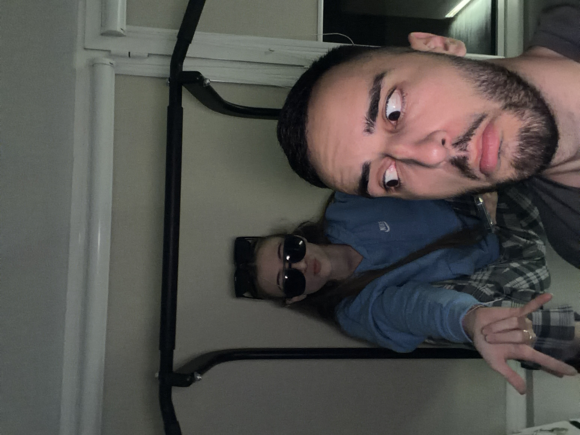Two people are visible in the image. One is a man with dark hair and a beard, making a surprised expression, with his eyes wide. The other person, presumably a woman, is wearing sunglasses and a blue sweater, with an arm making the "rock on" hand gesture. They are posed in what appears to be a narrow space, possibly a room. There is a black metal bar or frame in the foreground. The background shows light-colored walls and a window. The location is not discernible as Plovdiv, Bulgaria.