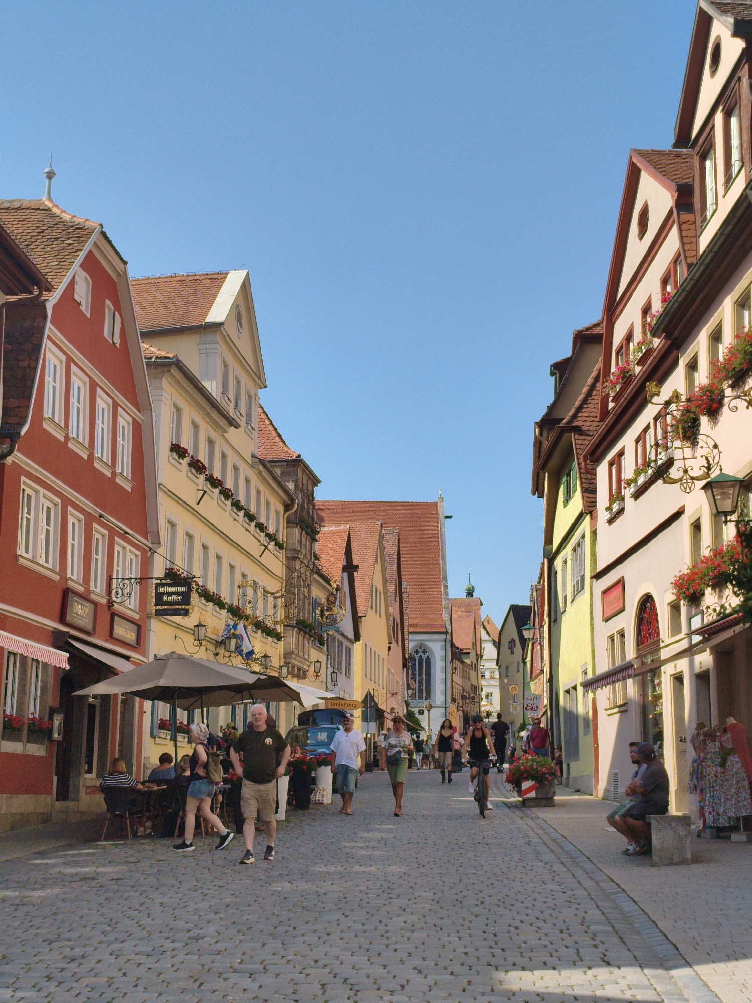 This image depicts a charming cobblestone street in Rothenburg ob der Tauber, Germany, on a clear, sunny day. The street is lined with traditional, colorful half-timbered buildings, adorned with window boxes overflowing with red flowers. Several people are walking along the street, while others are seated at outdoor tables of a cafe, enjoying the pleasant weather. The architecture and the presence of tourists suggest a historic and popular destination.