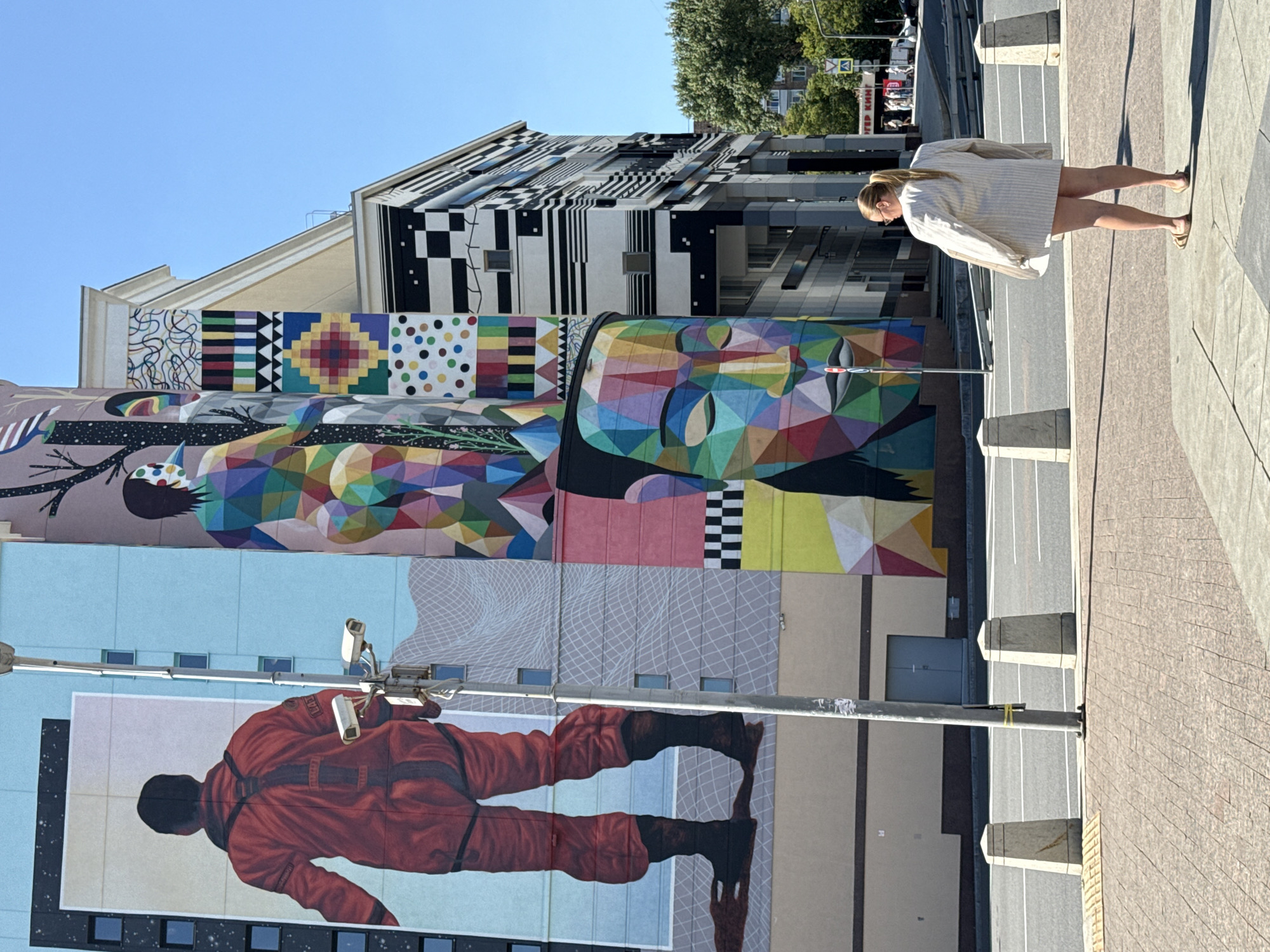 A woman with blonde hair tied in a ponytail stands facing away from the camera, wearing an oversized cream-colored pinstriped jacket and brown sandals. She is on a paved sidewalk in front of a large, multi-story building adorned with a vibrant mural. The mural features a figure in a red spacesuit on the left, a colorful abstract human figure in the center, and a large, geometric abstract face on the right. The building's facade also incorporates black and white geometric patterns. The scene is set on a sunny day in a city, with a clear blue sky above. The sidewalk is bordered by a street with a road and traffic barriers. In the background, trees and other buildings are visible, along with street signs and distant vehicles. The woman's pose suggests she is observing the artwork or the surrounding cityscape. The location is the Tverskaya Street area in Moscow, Russia.