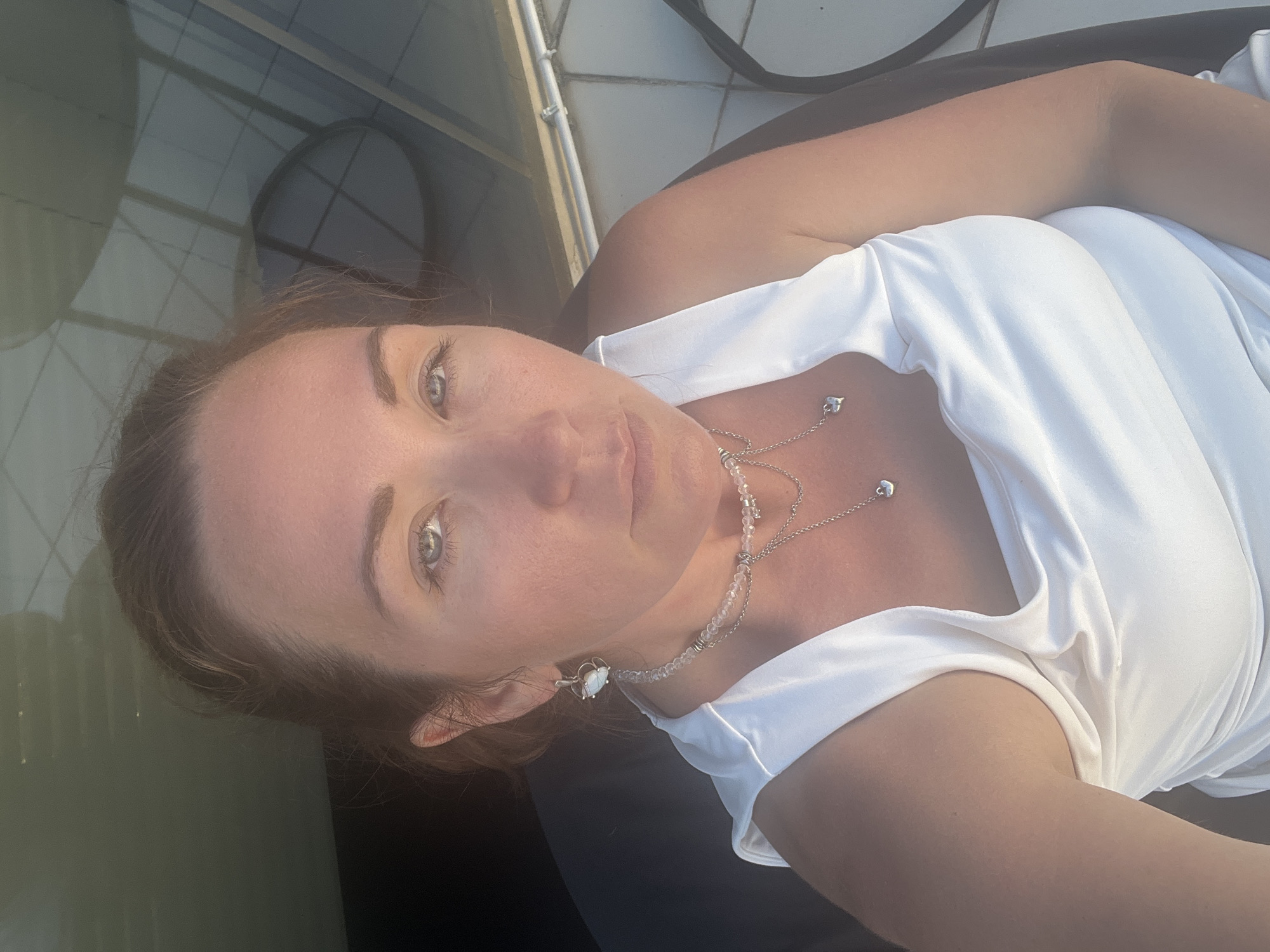 The image is a close-up, selfie-style portrait of a woman. She is positioned at an angle, with her head tilted upwards and her gaze directed towards the camera. Her fair skin is illuminated by natural light, highlighting her blue eyes and the subtle contours of her face. She has light brown hair that frames her face. She is wearing a white garment that appears to be a dress or top, with draped fabric visible. Around her neck, she wears a delicate silver necklace with what appear to be small beads and a charm.
The background suggests an outdoor setting, with a tiled surface and some shadows indicating a structure overhead, possibly a roof or pergola. There are hints of greenery and light reflecting on a surface, perhaps water or glass, on the left side of the image. The overall lighting suggests it is daytime, likely a sunny day, given the bright illumination and the shadows. The mood conveyed is relaxed and possibly serene. No visible text is present in the image. The metadata indicates the location as Sochi, Russia.
