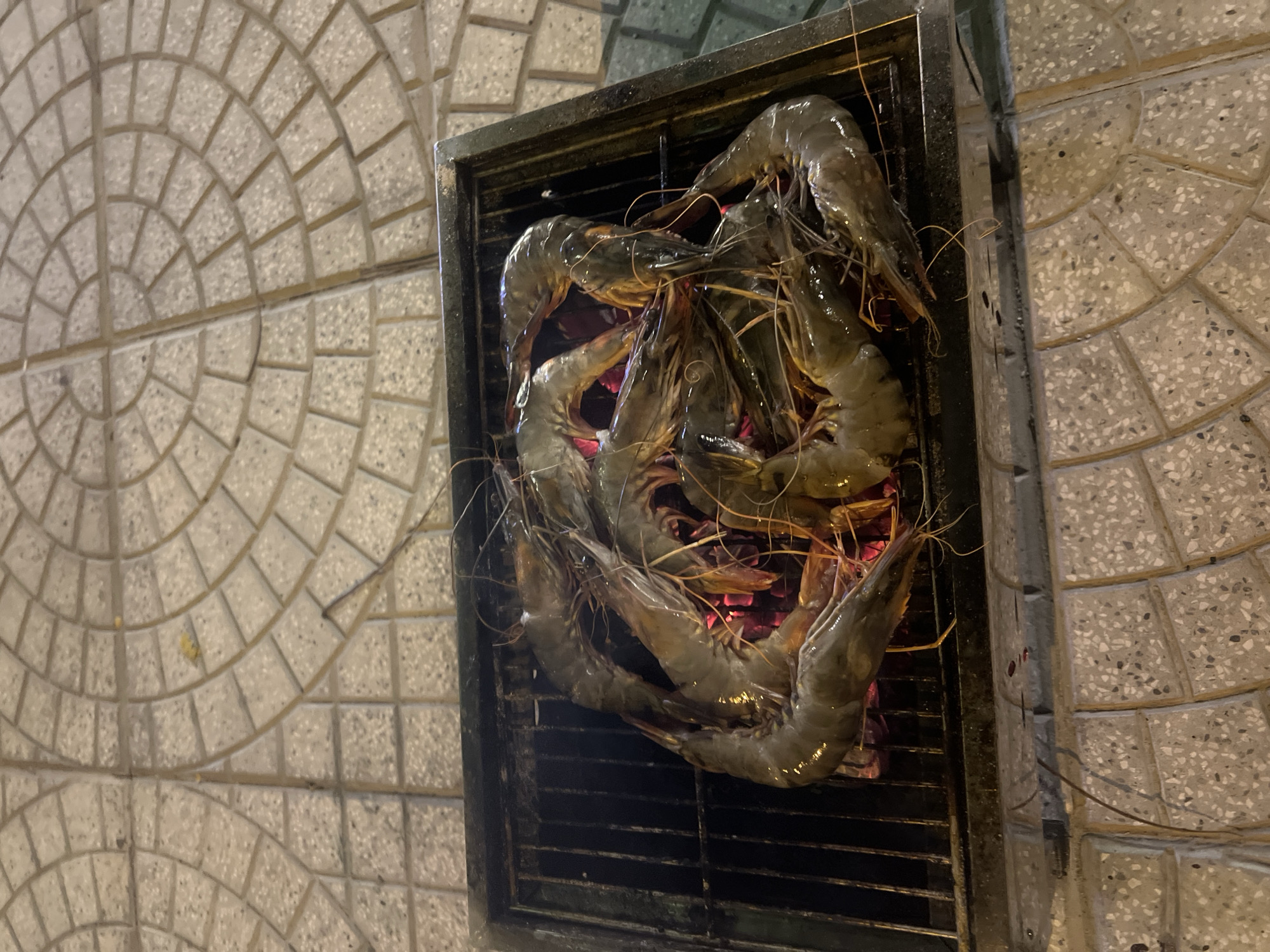 A close-up, high-angle shot shows several large, uncooked shrimp arranged on a metal grill over glowing red coals. The grill is situated outdoors on a paved walkway made of light-colored, speckled rectangular stones arranged in concentric arcs. The background is slightly blurred, but the textures of the paving stones are clearly visible. The lighting suggests it is nighttime. The scene appears to be in a public outdoor area, possibly a street or promenade, consistent with the urban environment of Đà Nẵng, Vietnam. There are no people visible in the frame.