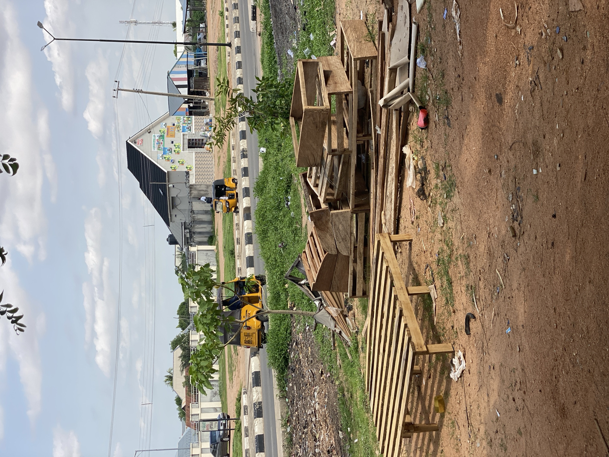 The image depicts a street scene in Jalingo, Nigeria, under a bright, cloudy sky, suggesting it is daytime.
In the foreground, a collection of wooden pallets and scraps are piled on the dusty ground. Some of these appear to be disassembled crates, while others are larger, unfinished wooden structures. Scattered on the ground are various pieces of debris, including plastic fragments, paper, and a single red shoe.
Adjacent to the pallets is a strip of green grass, and beyond that, a road with a black and white striped curb. On the road, a yellow motorized tricycle, commonly known as a "Keke Napep" or "Maruwa," is visible, with at least one person inside. Another similar tricycle is further down the road.
Behind the road, several buildings are visible. One prominent building has a dark, angled roof and a lighter facade adorned with various colorful advertisements and logos. Another building to its right is light-colored with a repeating pattern of windows. Further in the background, more buildings and structures can be seen, including what appears to be a telecommunications tower. Overhead, power lines stretch across the sky.
The overall impression is one of a casual, everyday scene in an urban or semi-urban environment in Nigeria, with elements of commerce, transportation, and the presence of discarded materials. The lighting suggests a sunny day, with scattered clouds.