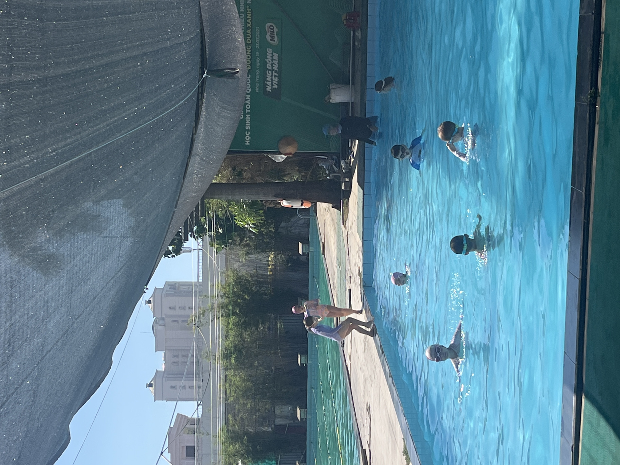 The image depicts an outdoor swimming pool in Nha Trang, Vietnam, under a large sunshade. Several individuals, primarily children, are engaged in swimming activities. Two young girls in swimsuits and caps are visible near the edge of the pool, one preparing to enter the water while the other stands on the pool deck. Several children are swimming in the pool, some wearing swim caps and goggles. In the background, there is a green wall with text, including the Milo logo, suggesting a sponsored event or venue. Trees and buildings are visible beyond the pool area. The water is a clear blue, and the overall atmosphere suggests a sunny day at a recreational facility.