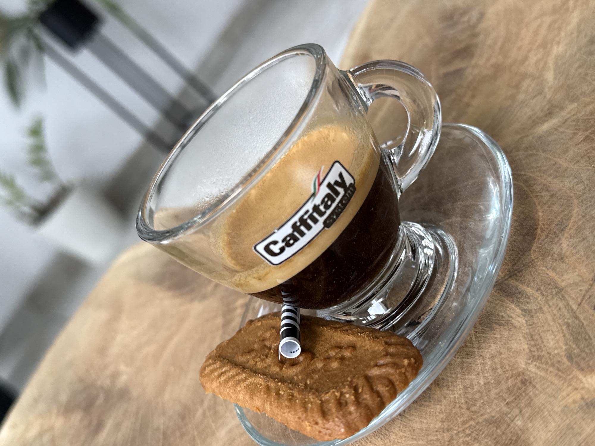 A tilted glass espresso cup with a "Caffitaly" label sits on a glass saucer atop a brown, textured surface. A cylindrical, black and white striped object appears to support the cup, positioned on a rectangular brown cookie on the saucer. The background shows blurred greenery and a structure composed of gray vertical and horizontal bars. The scene appears to be indoors. Location is not discernable from image content.
