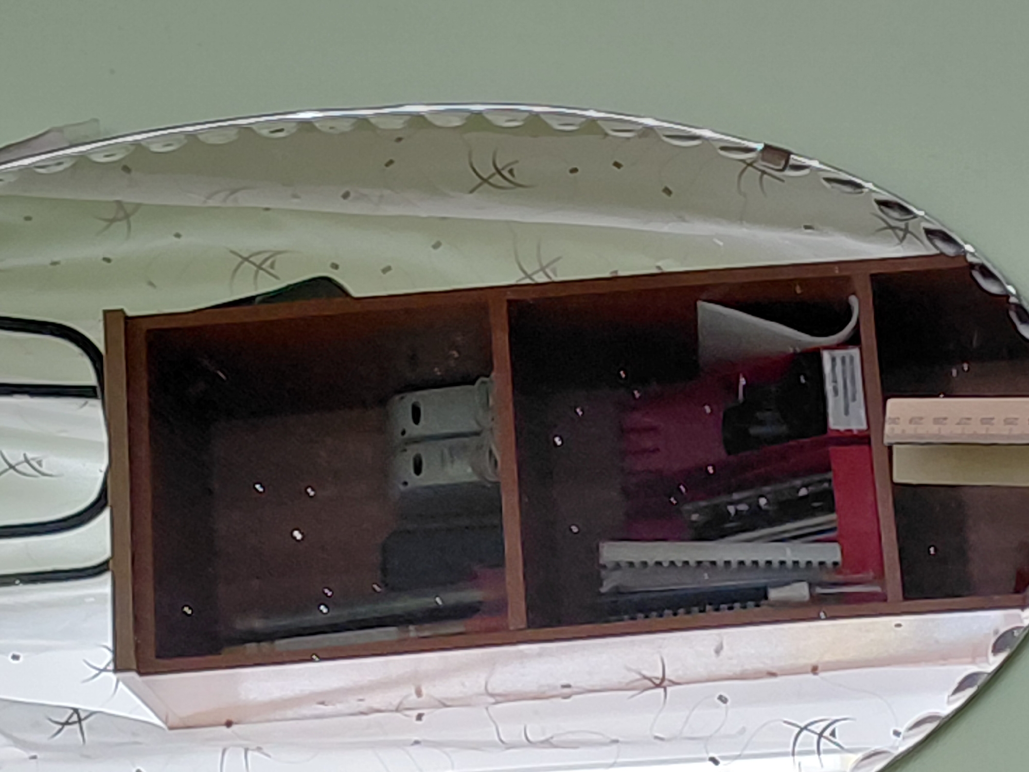 The image captures a reflection in a decorative, oval-shaped mirror with a scalloped, etched edge. The mirror is positioned against a plain, light green wall.
The reflection prominently features a wooden multi-compartment shelf or box. The shelf is divided into at least three visible sections, each containing various items. In the leftmost compartment, two light-colored, possibly metal, objects with oval cutouts are stacked, with other darker items below them. The central compartment contains a red-lined container holding several spiral-bound notebooks or binders and a white rectangular object with faint, undecipherable text, possibly "INFORMATION" or "ILLUSTRATIONS." The rightmost compartment holds a light wooden ruler with faint measurement markings and a stack of paper or a book. Small white specks, likely reflections of light sources, are visible within the compartments.
Above the wooden shelf in the reflection, a light-colored, possibly white or light green, curtain or fabric is visible. This fabric is patterned with abstract, dark, calligraphic-like lines and scattered small dots or squares.
No people are present, and no activity is taking place. The scene suggests an indoor setting, likely a study or office area in Nadym, Russia. The lighting is ambient, and no specific time of day can be determined.