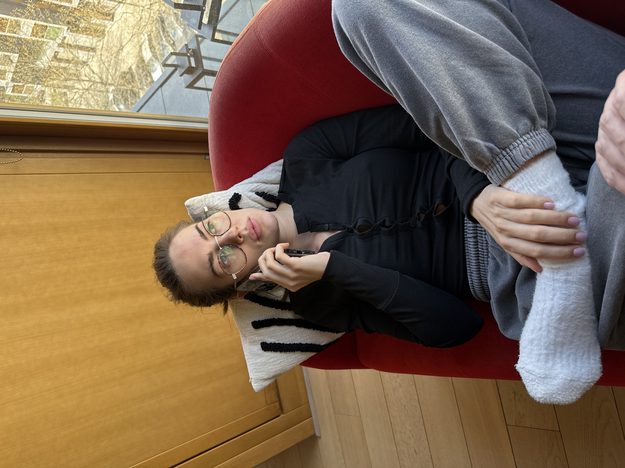 A young woman is captured in a medium shot, reclining sideways on a red armchair, seemingly engaged in a phone conversation. Her body is oriented horizontally across the frame, with her head resting on a white cushion featuring three black horizontal stripes. She wears round, wire-rimmed glasses, a dark long-sleeved top with a unique button or loop closure down the front, and light grey sweatpants. Her hair is pulled back, and she holds a dark-colored smartphone to her right ear with her right hand. Her left hand rests on her right leg. She is wearing textured white socks.
The setting appears to be indoors, likely a living room or study, with a large window visible in the upper part of the frame. Through the window, an urban landscape can be seen with reflections of buildings and bare trees, suggesting late autumn or winter. The room features a wooden floor and a light brown wooden window frame or wall panel. The overall lighting is bright, indicating daytime. The woman's expression is neutral and focused on her call. No visible text is present in the image. The scene is set in Warsaw, Poland.
