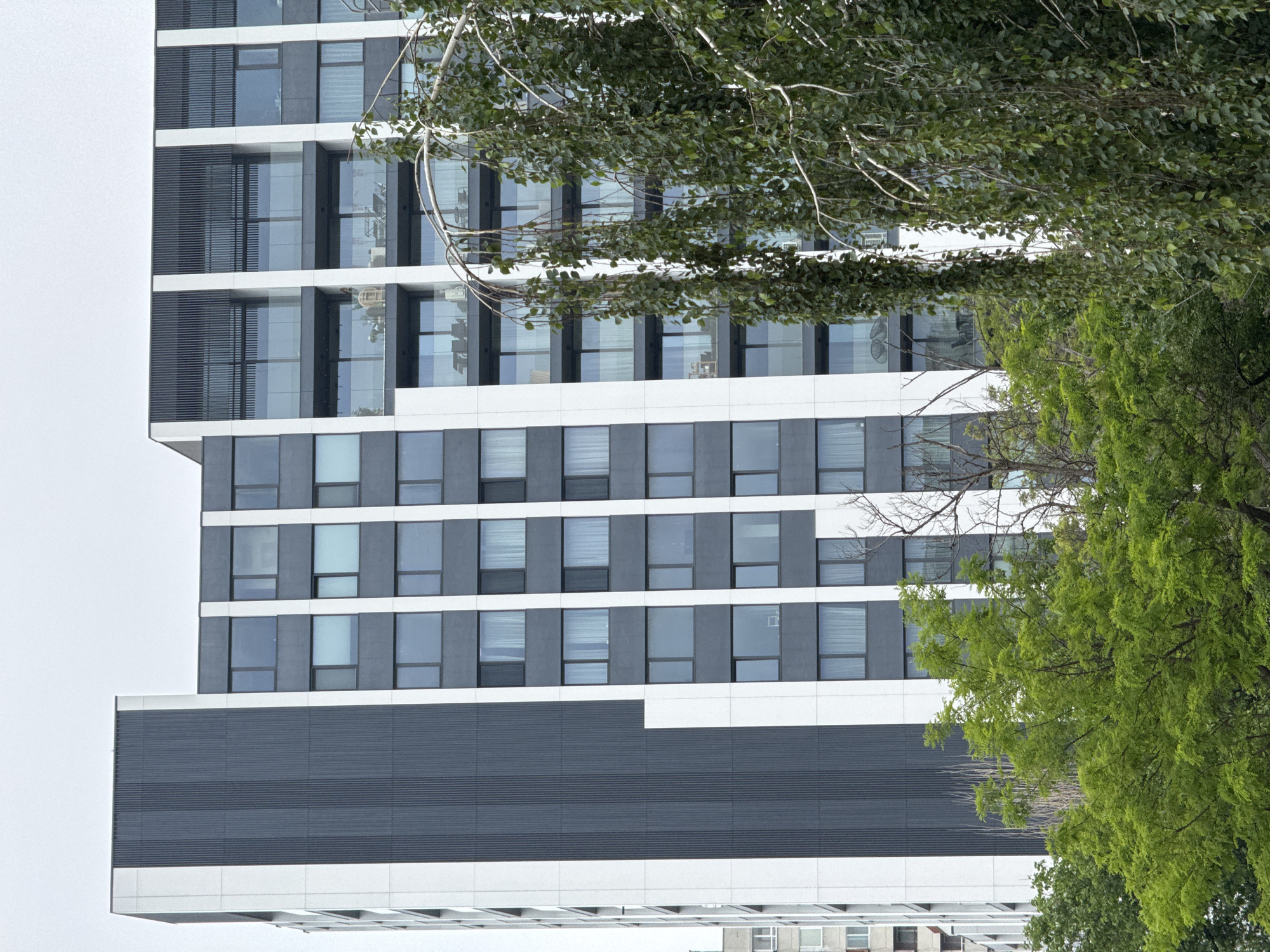 The image presents a close-up view of a modern, multi-story building partially obscured by vibrant green trees. The building exhibits contemporary architecture with strong horizontal lines, featuring alternating bands of white or light gray and dark gray panels. Numerous rectangular windows, mostly reflecting the bright sky or showing interior blinds, are arranged across the dark sections of the facade. A prominent dark gray cantilevered section, covered in horizontal cladding, extends from the lower half of the building.
Dense foliage from a tree with bright green leaves occupies the right side and foreground, with branches and leaves overlapping the building's facade. Another tree with sparser branches and lighter green leaves is visible amidst the denser foliage. The sky is uniformly bright white or light gray, suggesting an overcast day with soft, diffused light and an absence of strong shadows. No people or specific activities are depicted. The scene captures an urban setting in Varna, Bulgaria, combining natural elements with modern architecture. There is no visible text in the image.