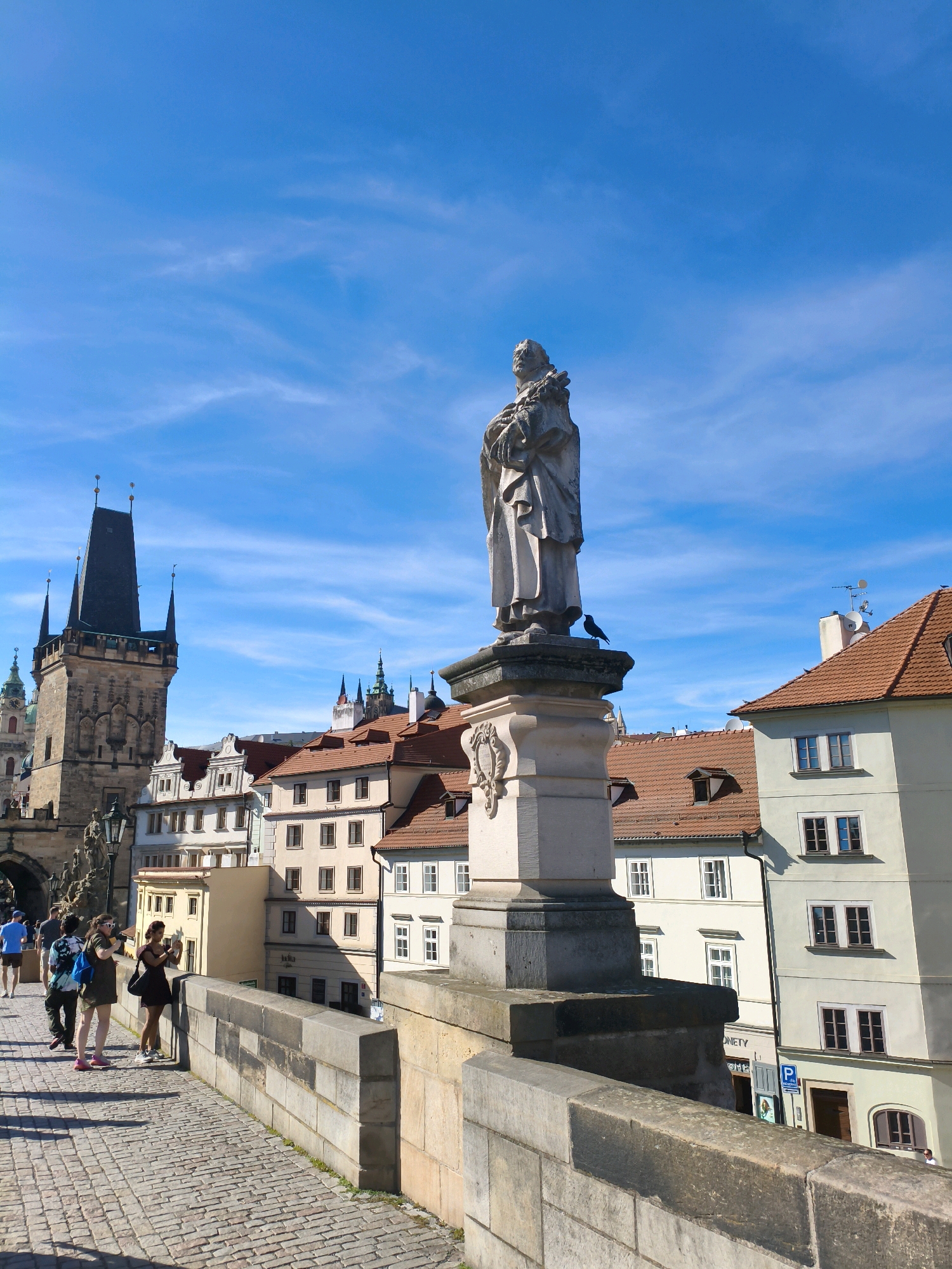 This is a vertical, eye-level, outdoor shot of the Charles Bridge in Prague, Czechia, on a bright, sunny day with a blue sky dotted with wispy white clouds. In the foreground, a stone statue of a man holding a book or scroll stands on a pedestal. A small black bird is perched on the pedestal to the right of the statue. The bridge is made of cobblestones, and a low, weathered stone wall runs along the right side of the frame. Several people are visible walking on the bridge. A group of three women is in the mid-ground, one of whom is taking a photo with her phone, and another is looking at her phone. Further in the background, a tall, dark tower with multiple spires dominates the left side of the frame, a characteristic feature of the Old Town Bridge Tower. Behind the tower and the statue, there are rows of historic buildings with terracotta roofs. The overall scene depicts a bustling, picturesque tourist destination.