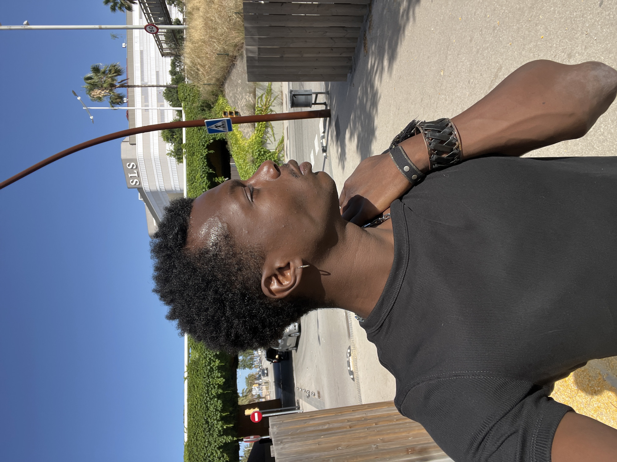 A young Black man is pictured in a medium shot, facing right, with his head turned slightly towards the camera. He has short, curly black hair and is wearing a black t-shirt. His left hand rests on his right shoulder, and he has two bracelets on his right wrist. The background suggests an outdoor urban environment with a clear blue sky. Behind him, a building with "SLS" signage is visible, along with palm trees and dry vegetation. A pedestrian crossing sign and a speed limit sign (50) are also present. A wooden fence runs along the right side of the frame. The man appears to be outdoors on a sunny day. This scene is located in Sant Adrià de Besòs, Spain.