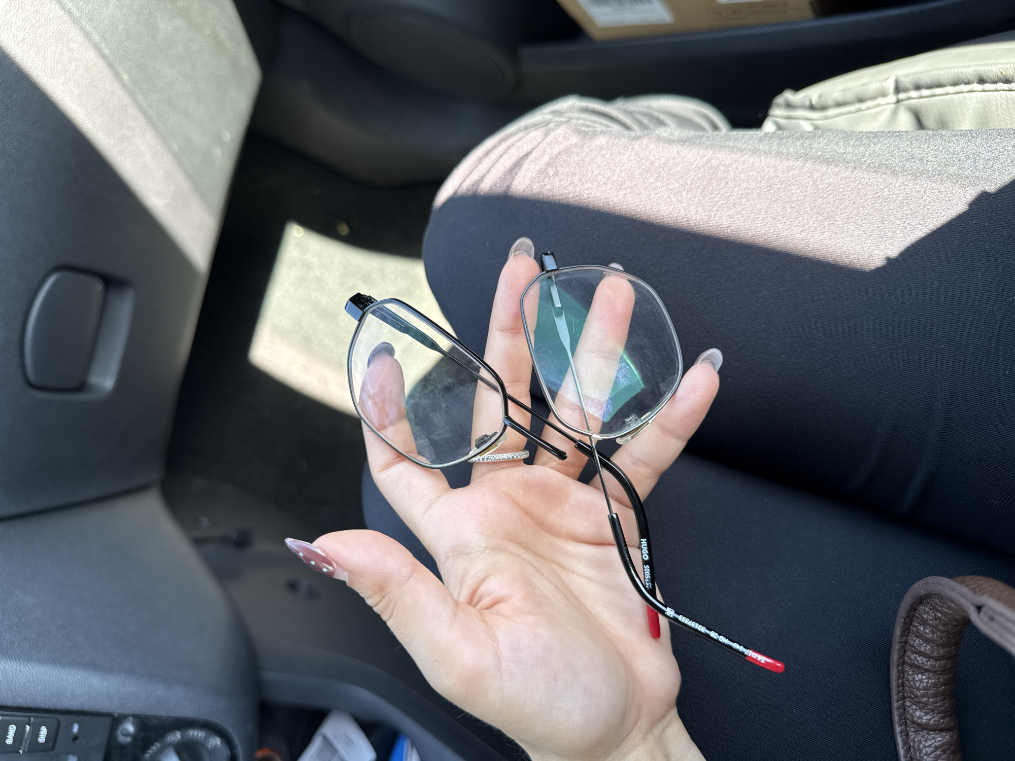 An interior view of a vehicle in Bruchköbel, Germany, shows a person's left hand holding a pair of black-framed Hugo 5005 prescription eyeglasses. The hand displays manicured nails, with one visible nail featuring white polka dots on a reddish-brown polish and another having solid red polish on its tip. A silver-toned ring adorned with small white stones is present on the ring finger. The eyeglasses feature clear, somewhat smudged lenses, and the temple arm clearly displays the branding "HUGO 5005."
The individual is wearing dark clothing on the left leg, while the right leg or adjacent car seat appears covered in a lighter grey, textured fabric. Bright sunlight streams into the vehicle, creating distinct bright streaks and shadows across the interior, including the person's leg and the car's headliner. Visible car interior elements on the left include a dark dashboard section with audio controls labeled "BAND" and "DISP," along with a rotary knob. A dark overhead compartment handle is visible on the upper right side of the frame. In the lower left, a brown, textured strap, likely from a bag, is partially visible. A cardboard box is partially visible in the bottom right corner of the frame, suggesting its presence inside or directly outside the vehicle.