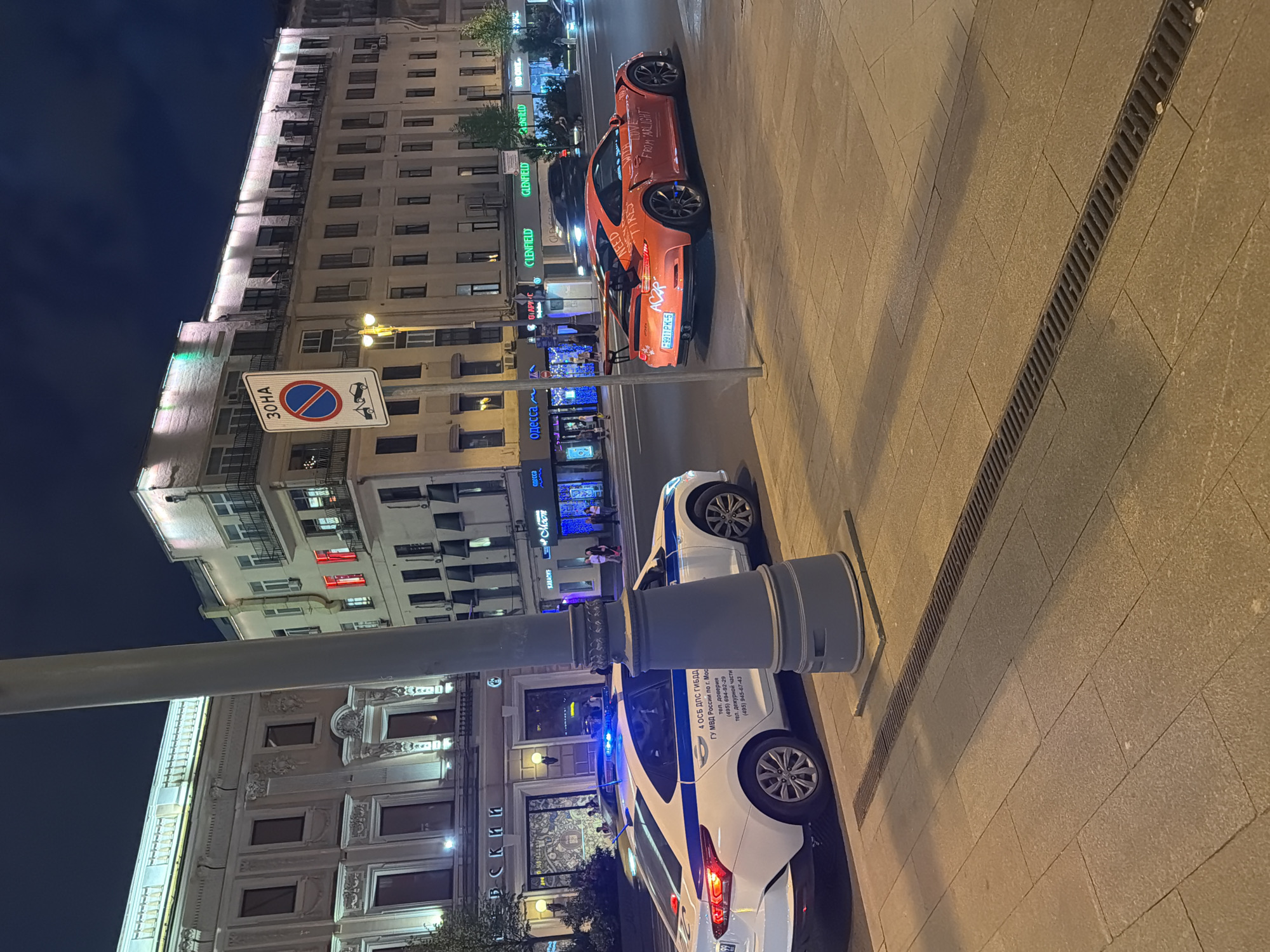 This night-time image captures a bustling city street in Moscow, Russia, with the pavement appearing damp, possibly from recent rain. In the foreground, a prominent street pole stands beside a white and blue Russian police car, whose blue emergency lights are flashing. Markings on the police car clearly state "4 ОСБ ДПС ГИБДД" and "ГУ МВД России по г. Мос," along with phone numbers: "тел. доверия (495) 694-92-29" and "тел. дежурной части (495) 945-67-43." A drainage grate runs along the tiled sidewalk next to it.
Further down the street, an orange sports car with a large rear spoiler is parked on the sidewalk. The car's side is adorned with graffiti-like text, including "ACZP," "NEED," "LOV," and "FROM ARLIGHT," suggesting the phrase "WE NEED LOVE FROM ARLIGHT." It has a Russian license plate "9911PK-5."
Multi-story buildings frame the street, featuring classic architectural details and numerous illuminated windows. The ground floors host various businesses with brightly lit signs displaying Cyrillic and English text, such as "GLENFIELD," "KARAOKE," "одесса" (Odessa), and "ПАРИС" (Paris). A white rectangular road sign is visible, featuring a red circle with a diagonal line, a tow truck icon, and the Cyrillic word "3OHA" (Zone) below it, indicating a "No Stopping" area. A few blurred pedestrians can be observed in the distance, contributing to the urban night atmosphere. The dark sky indicates the late hour.