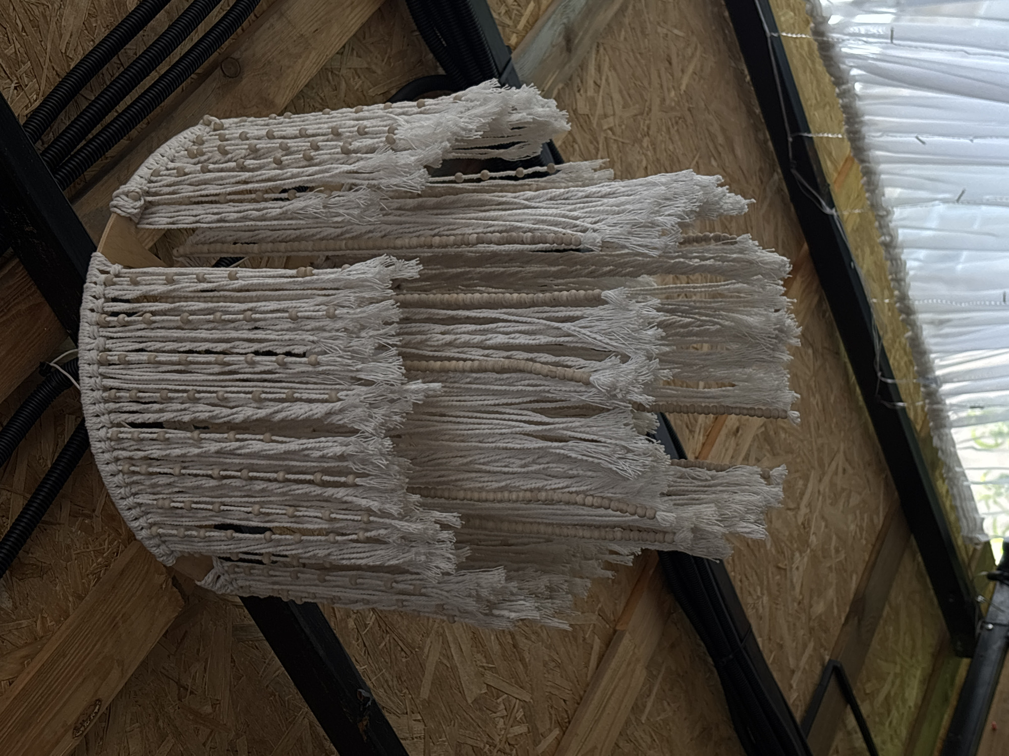 A two-tiered macrame chandelier with wooden beads hangs from a wooden ceiling with exposed beams and black metal supports. Black electrical conduit is visible on the ceiling. White curtains are visible in the lower portion of the image.