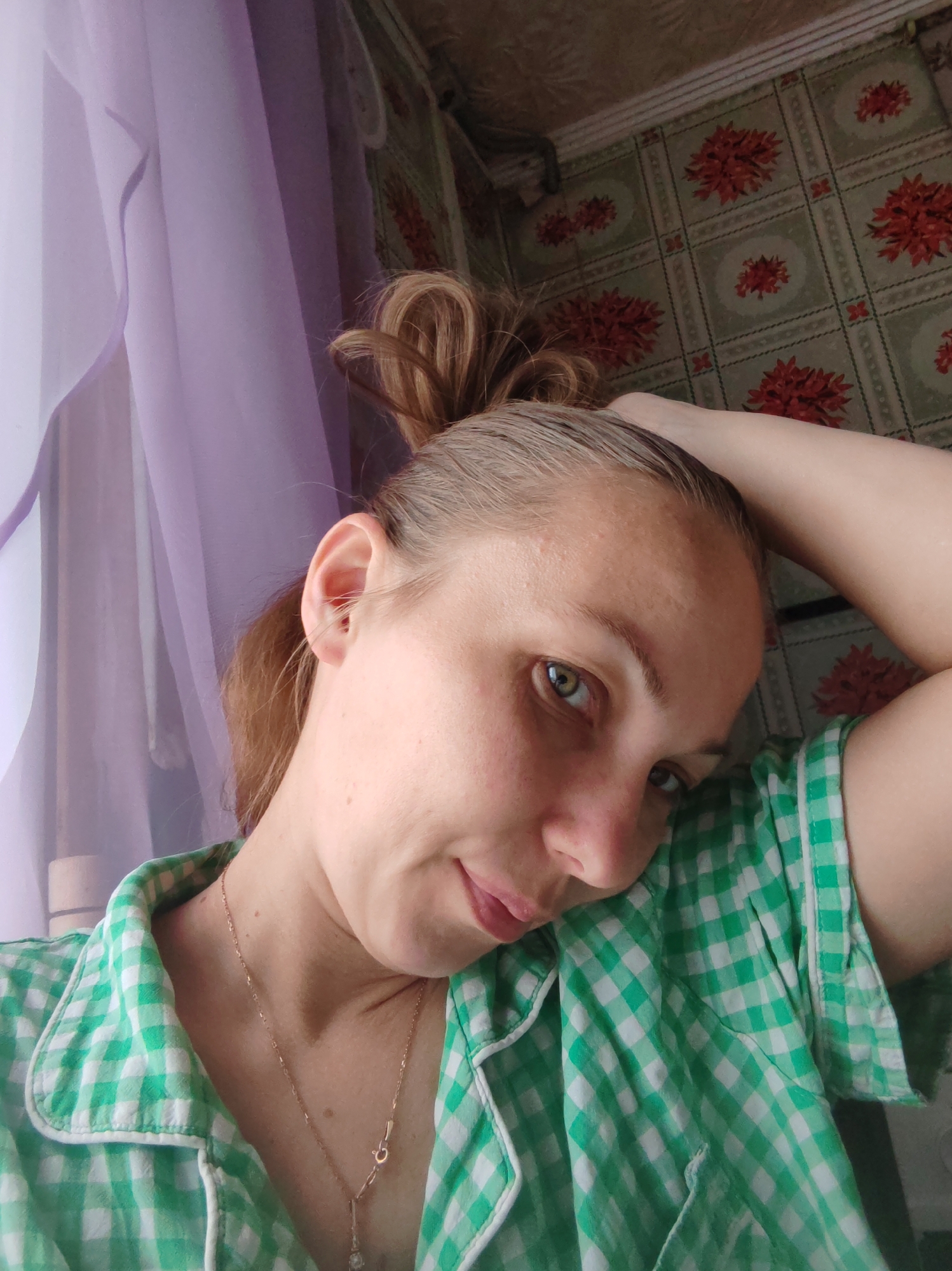A fair-skinned woman with blonde hair pulled into a messy bun is positioned in the foreground. She is wearing a green and white checkered shirt and a gold necklace. Her head is tilted, and she is looking slightly downward. Her right hand is raised and resting on the back of her head. Behind her, a wall is decorated with a patterned wallpaper featuring green and red floral designs. To the left of the woman, a light purple curtain hangs. The setting may be in an indoor environment. It is not possible to confirm the location in the city of Chukhloma, Russia, from this single image.