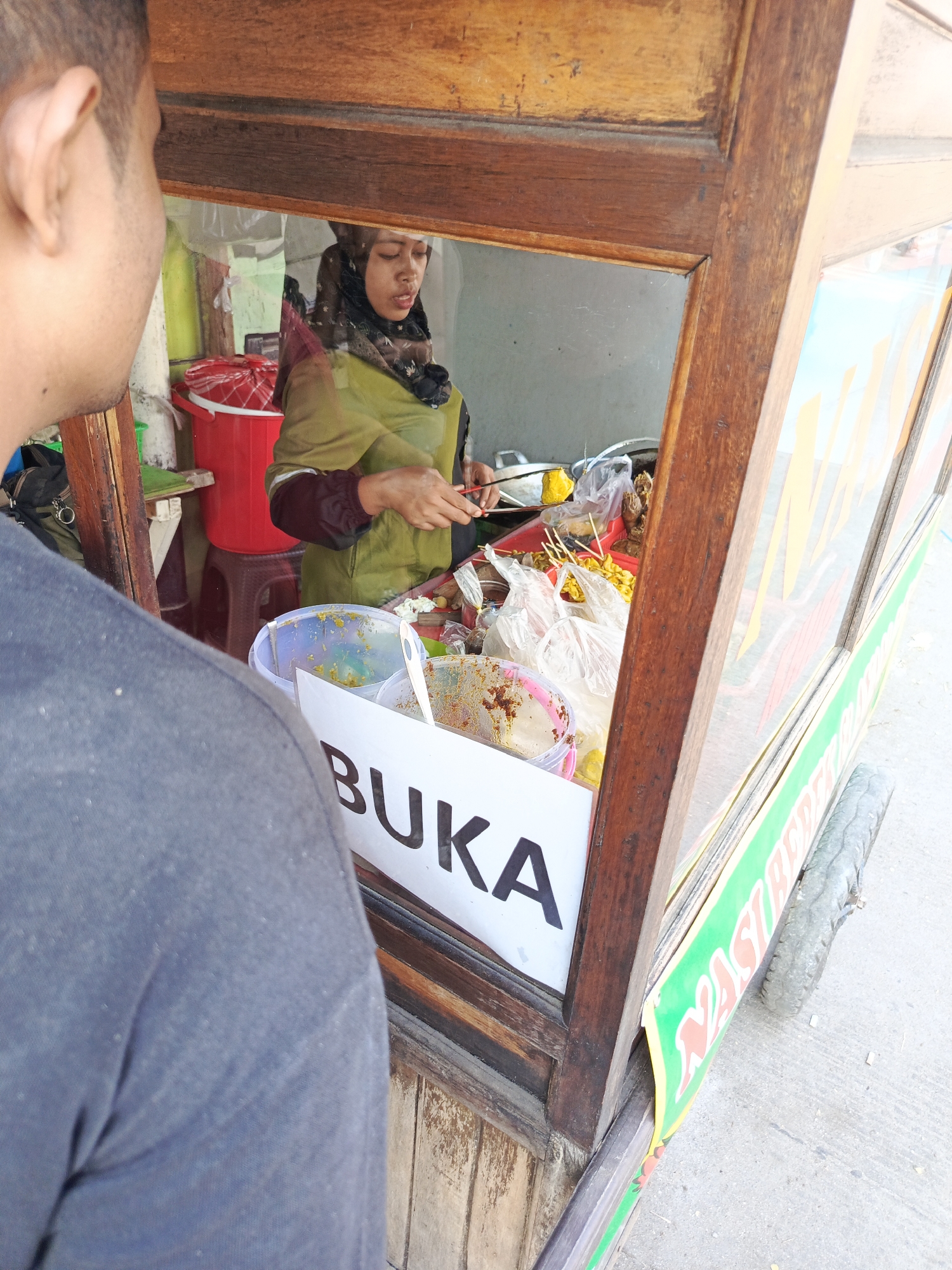 An over-the-shoulder shot from behind a person wearing a grey t-shirt focuses on a food cart vendor and her wares, viewed through the cart's wooden and glass window. The vendor, wearing a green headscarf and olive green attire, is preparing food, holding skewers of yellow food items in her right hand and a red utensil in her left. Inside the cart, several plastic containers hold various food items, including some that appear to be cooked meats and yellow skewers. A large red bucket is visible in the background. A white sign with the word "BUKA" (Indonesian for "open") is prominently displayed in the foreground. The exterior of the cart features a green banner with colorful lettering, possibly advertising food items such as "NASI GORENG" (Indonesian for "fried rice"). The cart is positioned outdoors, on a paved surface, with a wheel visible on the right side. The scene appears to be in a public area, likely a street or market in Tangerang, Indonesia, given the Indonesian language on the sign and banner.