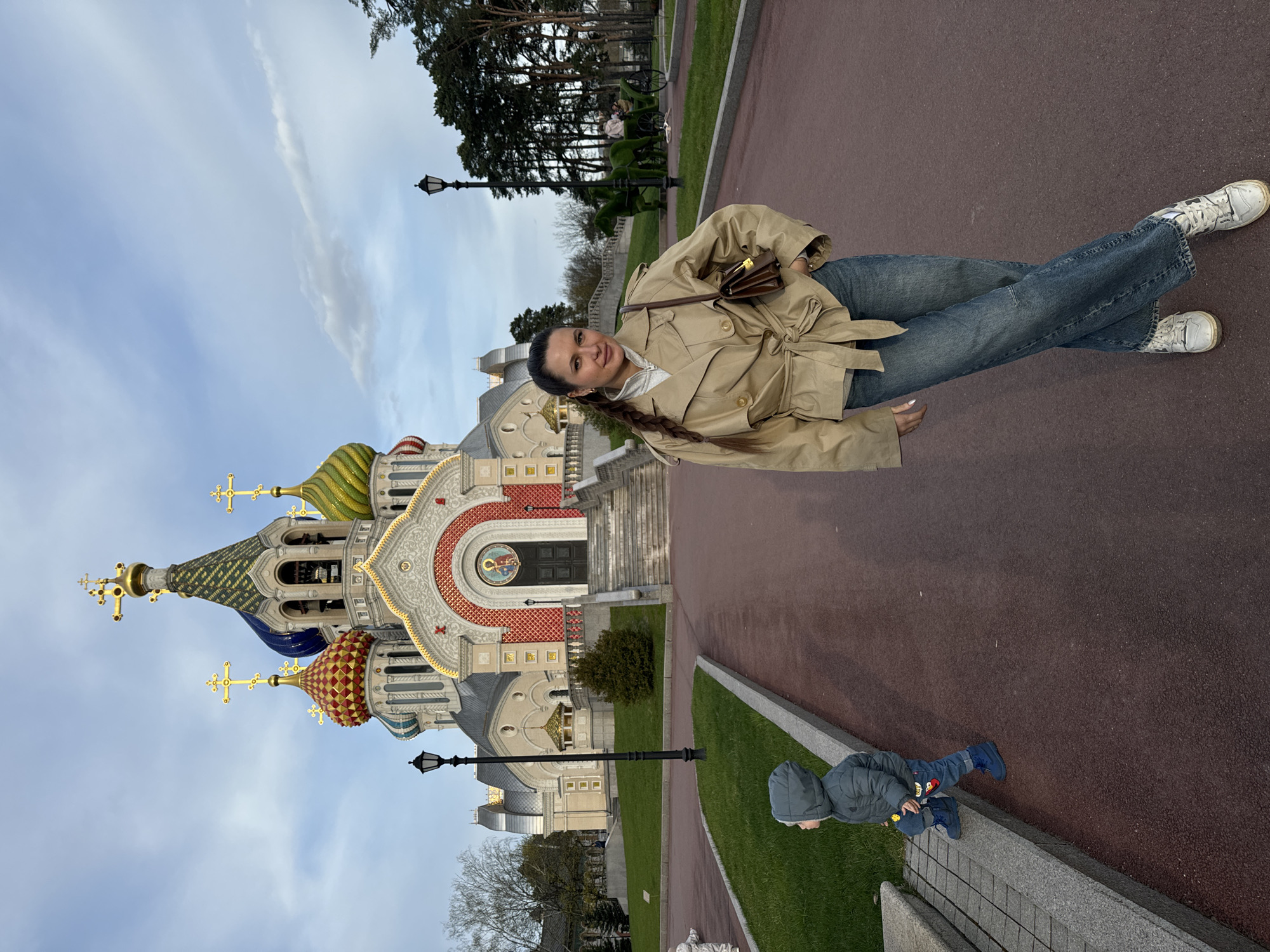 The image depicts a woman posing for a picture in front of the Church of All Saints, Moscow, Russia. The woman, who has long brown hair, wears blue jeans, a beige trench coat, and white sneakers. She is situated on a red paved area. Behind her is the elaborately decorated church with multiple colorful domes and golden crosses. The sky is visible behind the church, suggesting an overcast day. In the lower right corner, a child sits on a ledge near a grassy area. Several trees and lamp posts are also visible in the background.