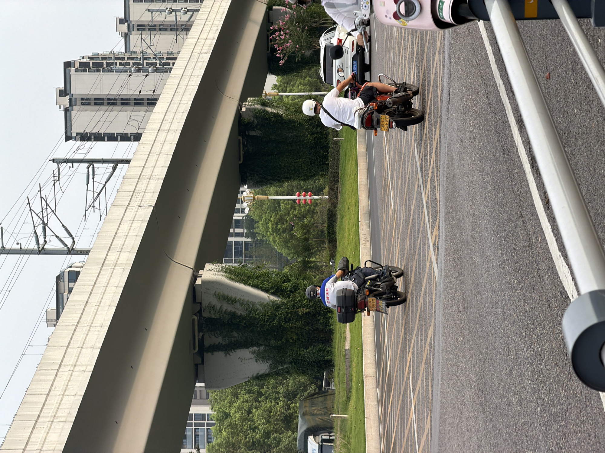 The image shows a view from a road in Jiangjin, China, looking towards a modern elevated structure, likely a highway or railway overpass. Several multi-story buildings are visible in the background, suggesting an urban or suburban setting. The foreground features a paved road with yellow and white lane markings. Three individuals on motorcycles are depicted. The first is a person wearing a blue shirt and a helmet, riding a black motorcycle with a storage box on the back. The second is a person wearing a white t-shirt and a helmet, riding an orange motorcycle with a white bag slung over their shoulder. The third motorcycle, a pink one with decorative elements, is partially visible on the right side of the frame, with a person on it wearing a white helmet. The overpass is supported by large concrete pillars covered in green vines. Trees and grass are present in the middle ground, bordering the road. The sky is overcast.