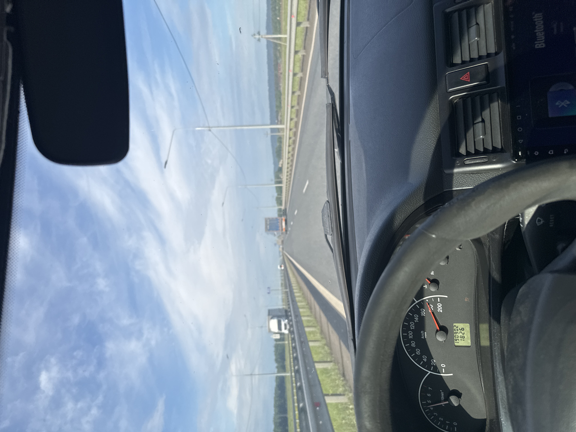 This media file captures a first-person perspective from the driver's seat of a car traveling on a multi-lane highway, likely near Усть-Сыны, Russia. The view looks forward through the windshield on a bright, clear day, with a blue sky sparsely dotted with wispy white clouds.
Inside the car, parts of the dashboard and steering wheel are visible, primarily on the right side of the frame. The speedometer indicates a speed of approximately 100 km/h, with "km/h" clearly legible. A digital display below the speedometer shows "190902" (likely the total odometer reading) and "1826" (possibly a trip meter). A tachometer marked "x100min-1" is also present. The dashboard features air vents, a hazard warning button with a red triangle symbol, and part of an infotainment system displaying "Bluetooth". A rearview mirror is partially visible in the upper left corner.
Outside, the highway stretches ahead with multiple lanes, bordered by guardrails and grassy verges. Utility poles or lamp posts line the road. Several other vehicles, including a white truck, are visible in the distance. The scene depicts a routine journey on an open road under good weather conditions.