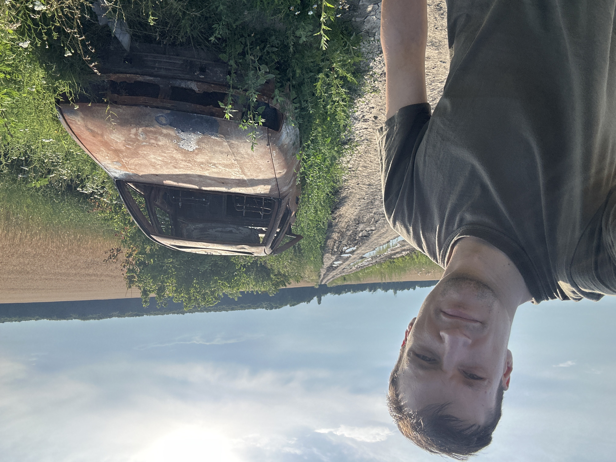 In Makiv, Ukraine, a man in a dark green t-shirt stands on a dirt road, facing the camera. To his right, a burnt-out car with its windows missing sits in overgrown grass. The road stretches into the distance, lined with fields and trees under a cloudy sky.