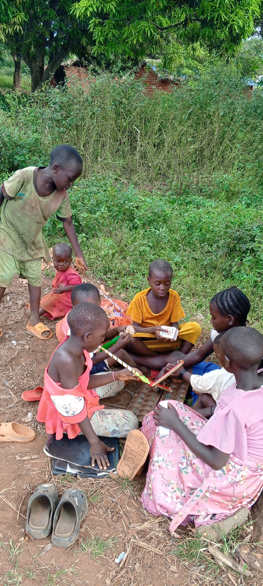 Seven children are gathered outdoors on a dirt ground with sparse grass, engaged in various forms of play. The scene is set in a rural area, with abundant green bushes and trees filling the background. Through the foliage, a brick structure with a rudimentary roof can be glimpsed, suggesting a dwelling.
In the foreground, a group of children sits on the ground. At the center, several children are focused on a traditional African board game, likely Mancala or Oware, which features a wooden board with depressions containing small playing pieces, possibly seeds or stones. A girl in a peach-colored top holds a long, striped stick, possibly part of another game or used to manipulate pieces. To her right, a girl in a yellow dress holds a stack of playing cards, looking down at them. Another girl in a pink floral dress sits facing the game, partially obscured.
Standing slightly to the left, a boy in a worn green t-shirt and shorts observes the seated children, looking downwards. A small child or toddler in a red outfit sits near his feet, also looking towards the activity.
The time of day appears to be daytime, with natural lighting. The children are dressed in simple, casual clothing. Several pairs of worn sandals and shoes are visible on the ground around them. One brown sandal sole clearly shows the letters "UCC," while a black sandal has "Skechers" partially visible on its side. The children appear engrossed and focused on their games. The overall impression is one of simple, communal play in a natural, rustic setting in Ngalula, Democratic Republic of the Congo.
