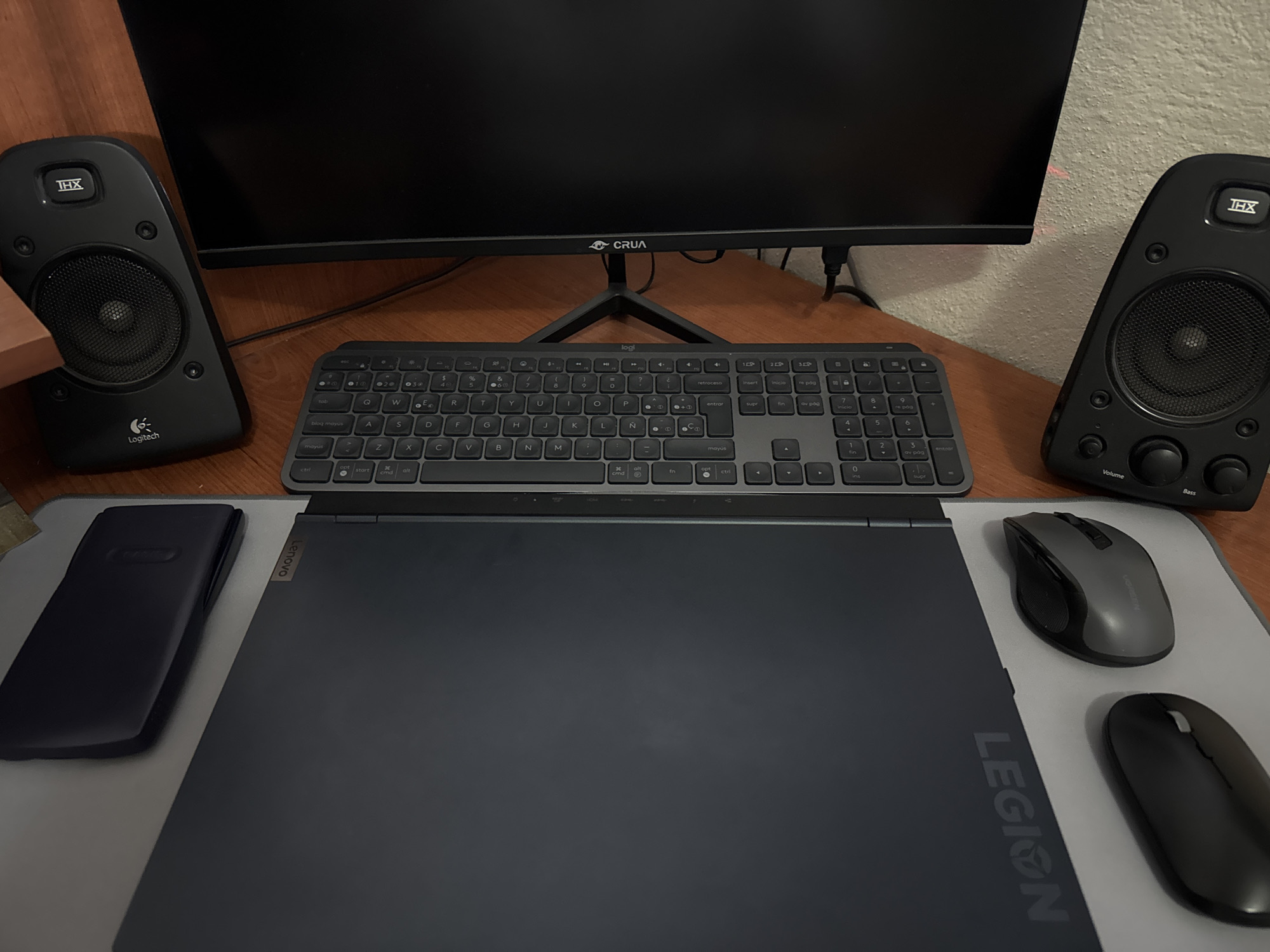 An indoor desk setup is captured from a slightly elevated perspective, showcasing a personal computing workspace. The primary subjects are various electronic devices and peripherals arranged on a light gray mousepad, which covers a wooden desk surface.
In the center, a black computer monitor, branded "CRUA," stands on its base. Flanking the monitor are two black Logitech THX speakers. The left speaker clearly displays "THX" at the top and "Logitech" at the bottom. The right speaker also features the "THX" logo and has two control knobs labeled "Volume" and "Bass."
Below the monitor sits a dark gray Logitech full-size keyboard with a number pad. Visible text on the keyboard includes "logi," "mayús," "retroceso," "inicio," "re pág," "supr," "fin," "av pág," "entrar," "ctrl," "start," "alt," "fn," and "cmd," suggesting a Spanish language layout, consistent with the location context of Puebla, Mexico.
In the foreground, a closed dark gray Lenovo Legion laptop rests on the mousepad. The "Lenovo" logo is visible on the hinge area, and "LEGION" is vertically printed along the right side of the laptop's lid. To the left of the laptop, a dark, rectangular wrist rest is positioned. To the right of the laptop, two computer mice are visible on the mousepad: one gray and one black.
The background reveals a light-colored, textured wall behind the monitor. No human activity or specific event is taking place; the image presents a stationary, organized workstation. The lighting appears to be indoor, without any discernible time of day or weather conditions.