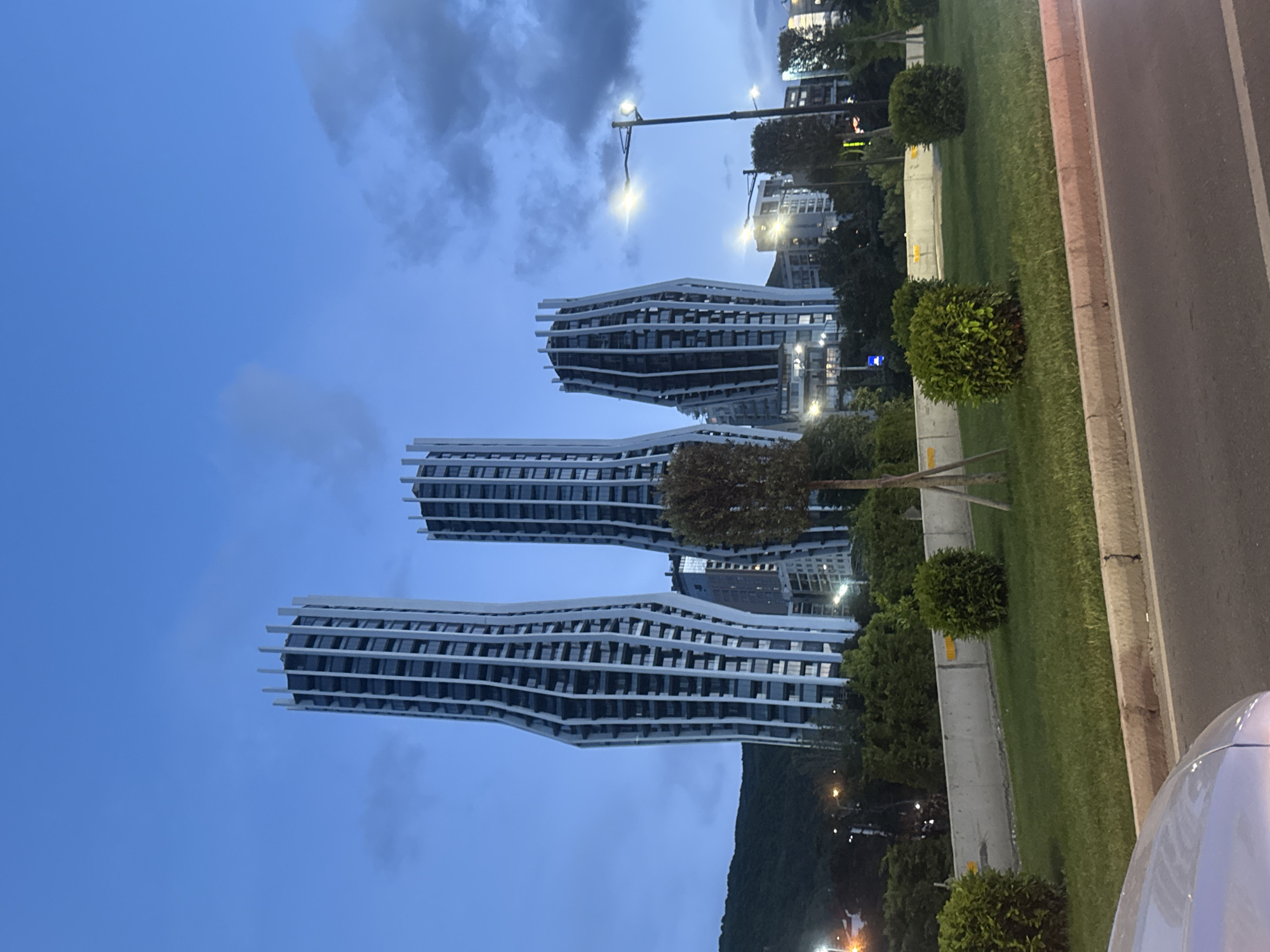 The image shows three modern, wavy-designed buildings in Tbilisi, Georgia. The buildings are white with dark windows, and they appear to be residential towers. The sky is a dusky blue with scattered clouds. A street and green space are visible in the foreground, with manicured bushes and trees separating the buildings from the road. A streetlight is on the right side of the image. A small portion of a white car is visible in the lower right corner. The buildings are positioned vertically in the frame.