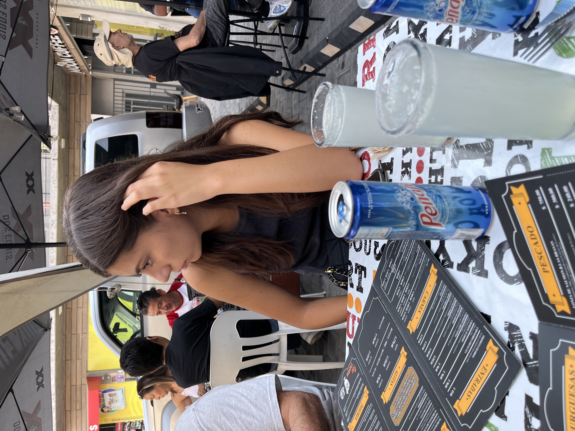 Here is a detailed description of the image:
The photograph captures an outdoor dining scene, likely a restaurant or cafe, in San Francisco Totimehuacán, Mexico. A young woman with long, dark brown hair is seated at a table. She is wearing a black top and has her hand near her face, possibly adjusting her hair.
On the table, there are various items: a blue can of "Peñafiel" mineral water, two tall glasses with a light-colored beverage (possibly lemonade), and a menu with sections labeled "Burguesas", "Pescado", "Carnes", "Entradas", "Alitas", and "Postres" (Burgers, Fish, Meats, Entrees, Wings, and Desserts).
Other people are visible in the background at adjacent tables. A man is wearing a white shirt with a red and white jacket. Another man is wearing a black shirt and is seated at a table. There is a portion of a white plastic chair also visible. The background shows a glimpse of the town, with a white delivery truck and buildings.
An umbrella provides shade above the table.