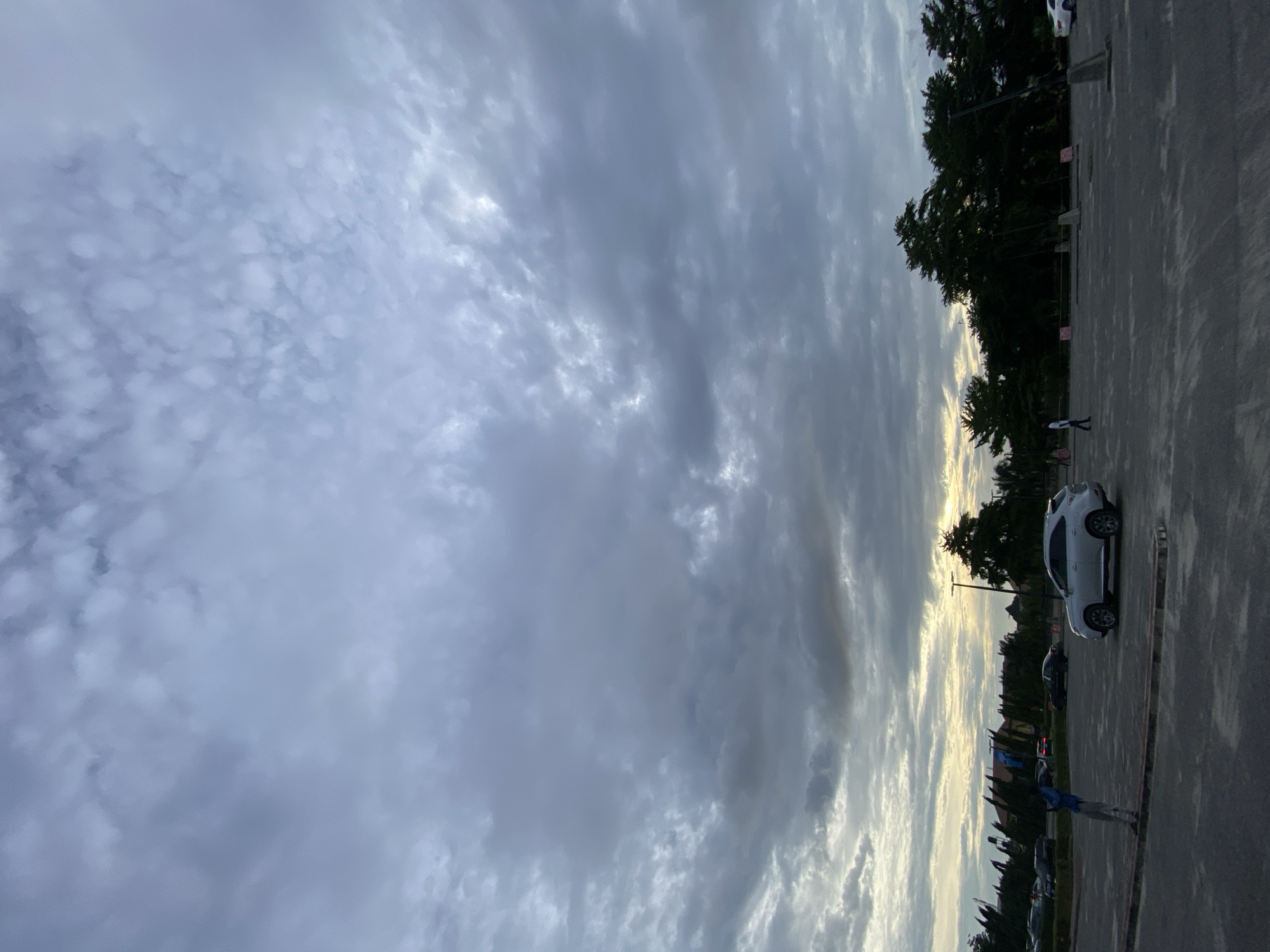 The image shows a cloudy sky above what appears to be a parking lot. The scene is likely at dusk or dawn, given the lighting. The parking lot is sparsely populated with cars. Trees and a silhouette of buildings are visible in the background. One person stands near parked cars in the foreground. The location is possibly Sangotedo, Nigeria, based on the request.