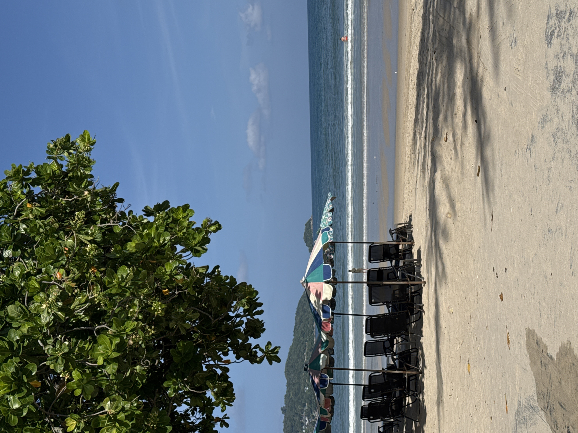 In Patong, Thailand, a tranquil beach scene unfolds under a clear blue sky. The foreground features a row of empty black lounge chairs arranged neatly under colorful beach umbrellas. The sand is smooth with some shadows cast from overhead foliage, suggesting a partially shaded area. Beyond the chairs, gentle waves lap at the shore of the Andaman Sea. In the distance, a lone individual can be seen wading in the water. Lush green hills form the backdrop, indicating a coastal setting within the tropical landscape of Patong. The overall impression is one of peaceful relaxation on a sunny day.