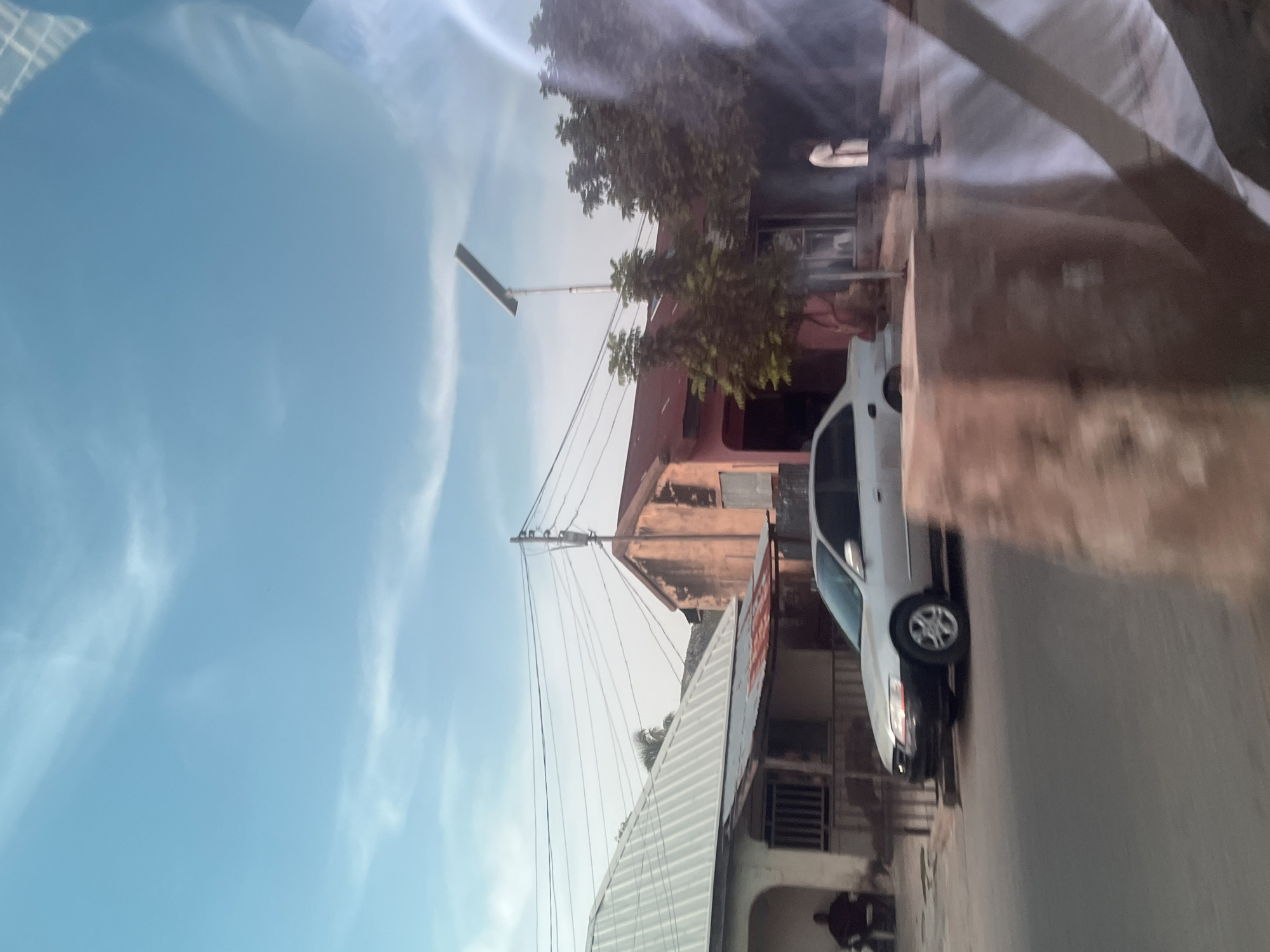 The image depicts a street scene in Jalingo, Nigeria, viewed from a slightly elevated perspective, possibly from a vehicle. The sky is a clear, pale blue with wispy white clouds, suggesting it is daytime, likely morning or afternoon. The orientation of the image is rotated, so the street appears vertical and the sky horizontal.
The scene includes several buildings of varying architectural styles and states of repair. One building, painted in a faded reddish-brown, has multiple windows and a dilapidated appearance. Another building features a white, sloped corrugated metal roof. Power lines are strung across the scene, supported by poles, indicating an active, inhabited area.
A white sedan is parked or driving on the street, its front passenger side wheel clearly visible. The car appears to be in good condition. In the foreground, a large white tarp or sheet is draped over something, casting shadows and adding a sense of texture to the image.
There are faint hints of people in the scene, with one figure visible in silhouette near the bottom left, near the white tarp. Another figure can be seen on a balcony or upper floor of one of the buildings. The overall impression is of a typical urban or semi-urban environment in Nigeria, with a blend of residential and commercial structures, infrastructure, and daily life. The lighting suggests a bright day, and the absence of strong shadows implies the sun is not directly overhead. No visible text is discernible in the image.