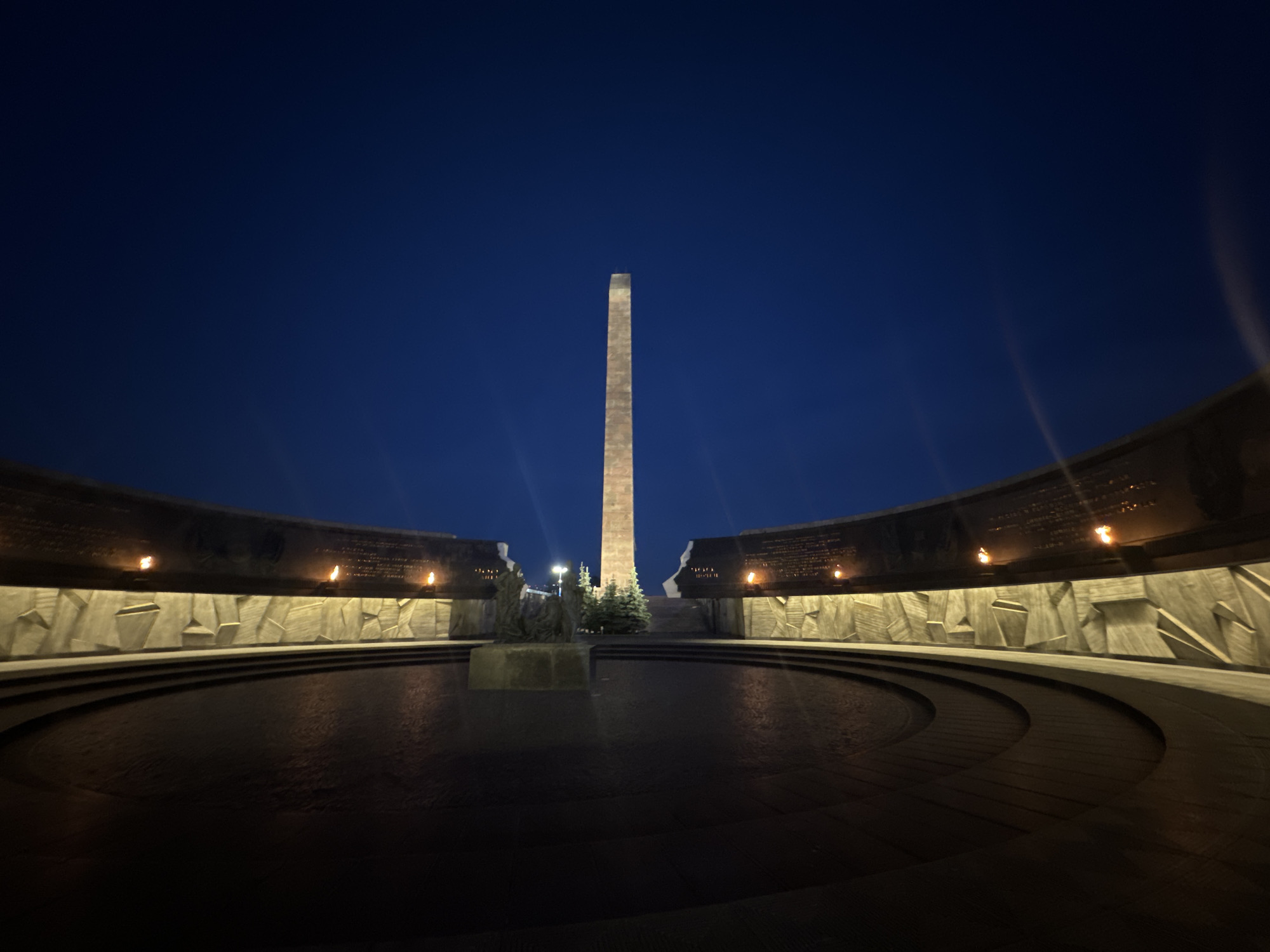 This nocturnal image displays the Monument to the Heroic Defenders of Leningrad at Victory Square (Ploshchad Pobedy) in Saint Petersburg, Russia. A tall, slender obelisk, illuminated in a light tone, stands centrally against a deep blue sky. It is flanked by two large, curving walls of concrete or stone, which form a semi-circular perimeter. The lower portions of these walls are adorned with geometric, angular relief patterns. Along the upper edge of the curved walls, multiple small, bright orange eternal flames burn. Subtle inscriptions or reliefs are visible on the darker, upper sections of the walls above the flames. In the foreground, a large, dark, circular basin, likely filled with water, occupies the ground, exhibiting faint reflections. A dark, multi-figure sculpture group, depicting human forms, is positioned on a pedestal within this basin. Scattered evergreen trees are present at the base of the obelisk and behind the central sculpture. The entire memorial complex is illuminated by artificial light sources, creating distinct light streaks in the lower frame. No individuals are present in the scene.
