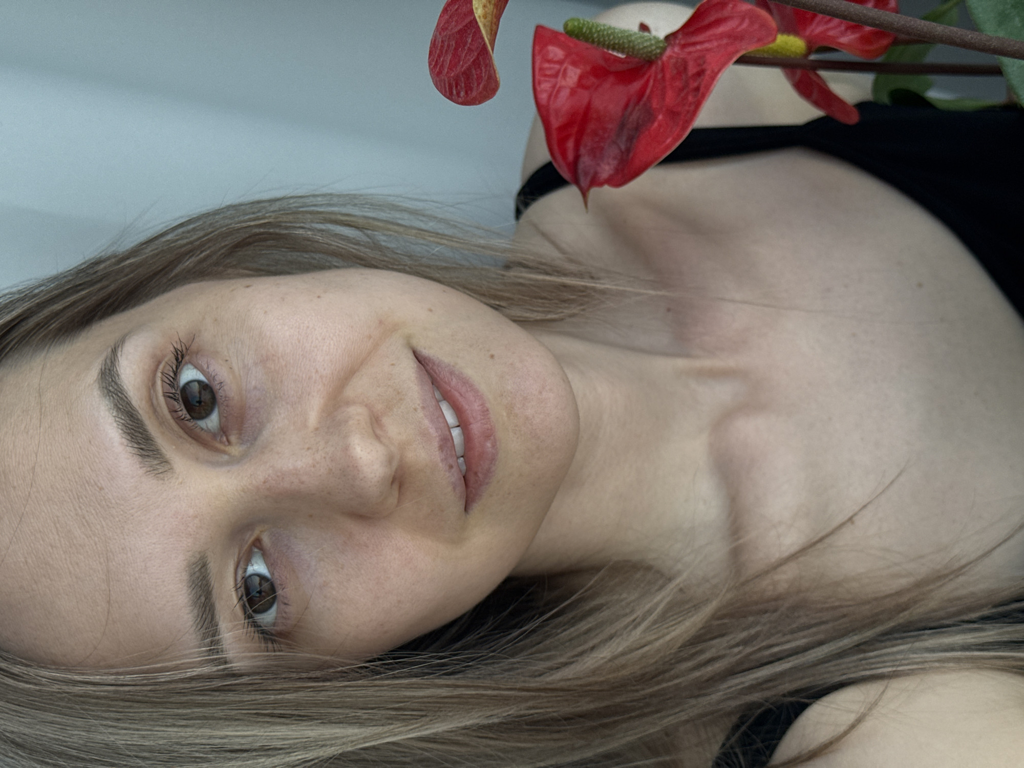 A woman with fair skin and light brown hair is lying down, facing upwards. She has brown eyes and a slight smile. A red anthurium flower is positioned near her shoulder. She is wearing a black top. It is not possible to determine the location as Chaikovsky, Russia from the image alone.