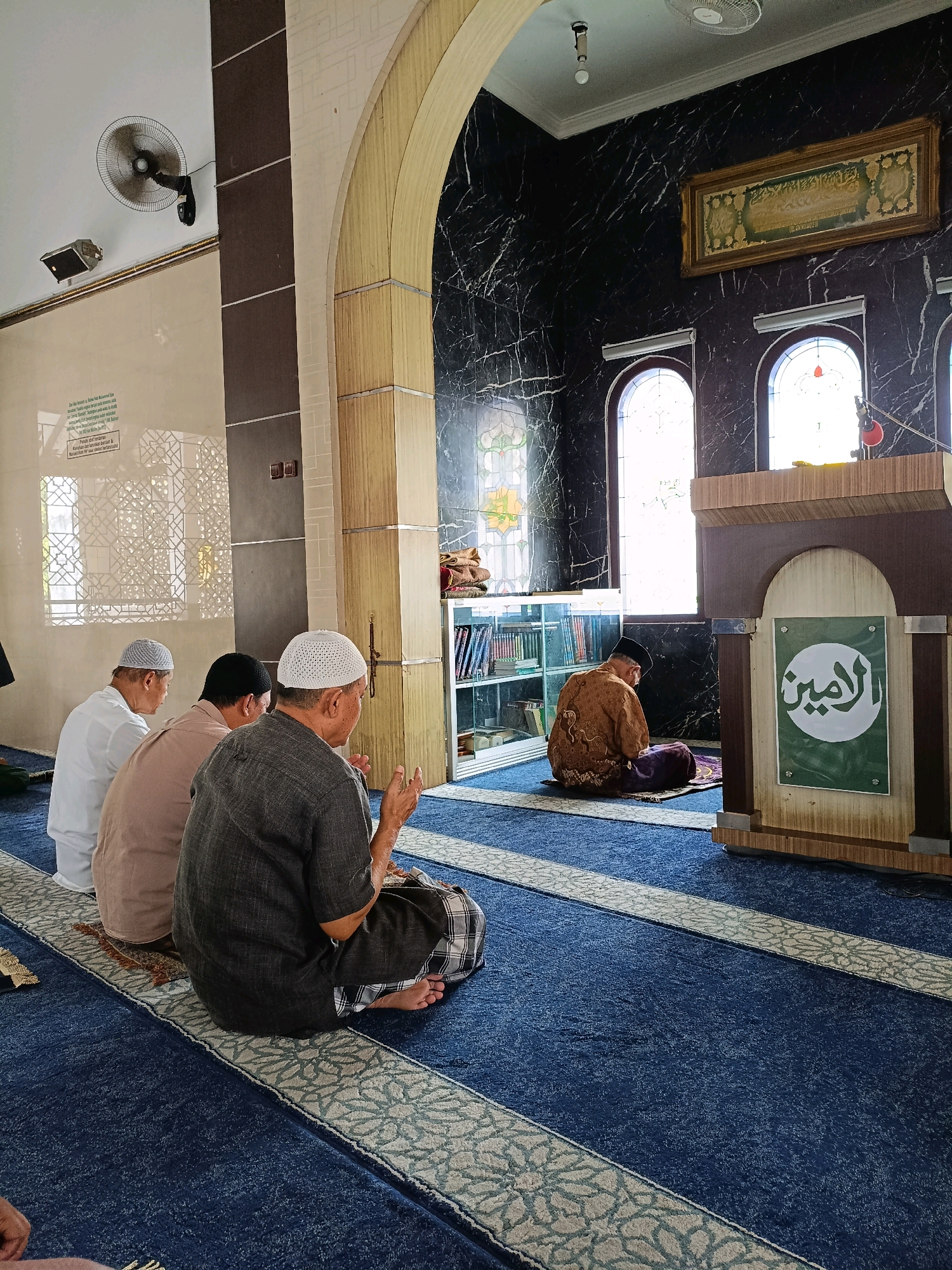 The image depicts the interior of a mosque in Maguwoharjo, Indonesia, during prayer. Four Muslim men are shown seated on a blue carpet with a patterned runner. Three men are in the foreground, facing away from the camera and towards what appears to be the direction of prayer. One man in the far left is wearing a white shirt and cap, the man next to him a brown shirt and cap, and the man in the middle foreground a dark grey shirt and white cap. The fourth man is further back on the right, wearing a brown patterned robe and a black cap, also facing away and seated on a prayer mat. The environment features arched doorways, dark marbled walls, and a wooden pulpit with a green sign displaying Arabic calligraphy. A ceiling fan is visible in the upper left of the image. The lighting suggests it is daytime, with light filtering in from windows.