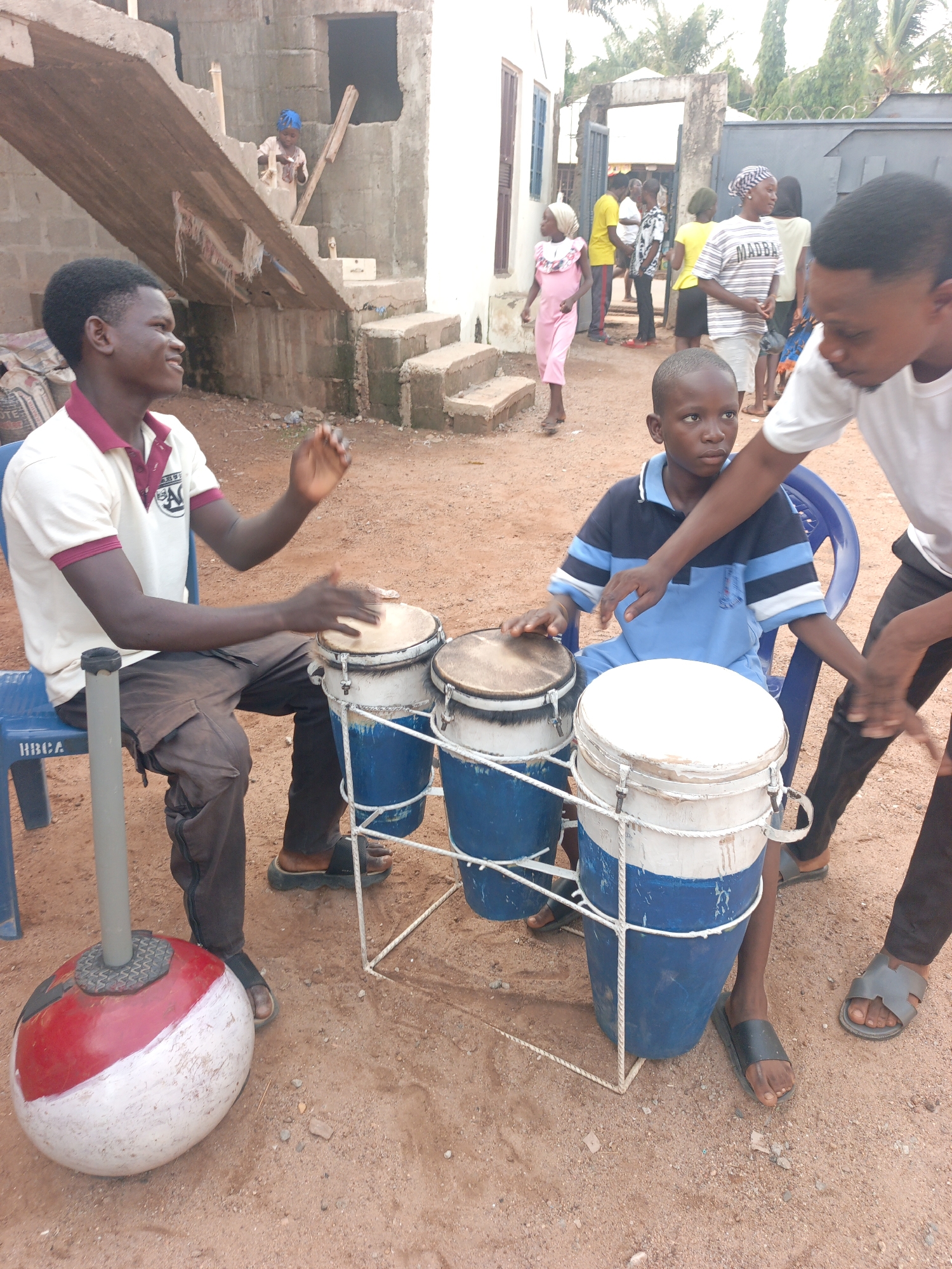 This image captures an outdoor scene in what appears to be a semi-urban or rural setting, likely in Akwanga, Nigeria, given the provided context. The ground is dirt, and unfinished concrete buildings are visible in the background, one with exposed rebar and a partial staircase.
In the foreground, three males are engaged with a set of drums. To the left, a young man, wearing a cream polo shirt with maroon trim and dark pants, sits on a blue plastic chair (partially obscuring text "HBCA") and plays a small drum. He is smiling and appears to be enjoying the activity, with his hands blurred from movement. Next to him, a young boy, wearing a blue and white striped polo shirt, sits on another blue plastic chair, focused on the drums. He has his hands on the drum heads, suggesting he is either learning or participating. Standing to the right of the boy is another young man in a white t-shirt and dark pants, leaning over and seemingly guiding or teaching the boy how to play the drums, with his hand near the boy's.
The drum set consists of three drums, constructed from blue and white barrels or containers, with rope detailing and what appear to be animal skin or synthetic heads. A red and white spherical object, possibly a buoy or a decorative item, is positioned on the ground to the left of the drums, with a gray pole emerging from it.
In the midground and background, several other people are visible. On an incomplete concrete staircase in the background, a young child with a blue head covering stands. Further back, near a gate and another building, a group of people, including children and adults, are standing or walking. One person wears a striped t-shirt with the word "MADBA" visible. The overall lighting suggests it is daytime and likely sunny. The expressions of the main subjects convey engagement and a positive, communal atmosphere.