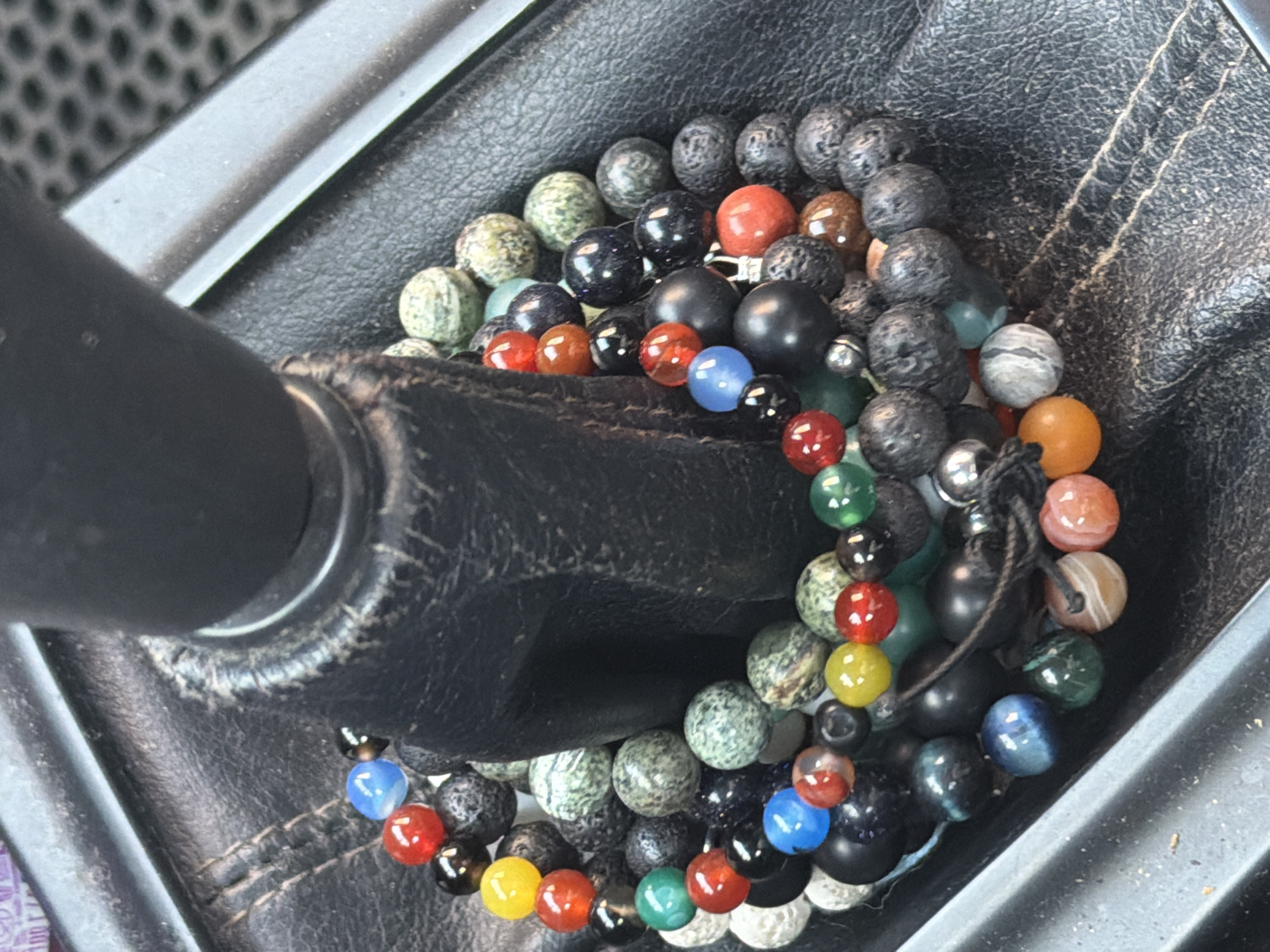 A close-up shot shows multiple beaded bracelets encircling a black leather gear shift boot. The bracelets consist of beads in various colors and textures, including red, blue, green, yellow, black, orange, and mottled gray-green. The environment includes the surrounding black plastic and metallic components of the vehicle's interior. No people are visible. The location cannot be identified as Izhevsk, Russia without additional information.