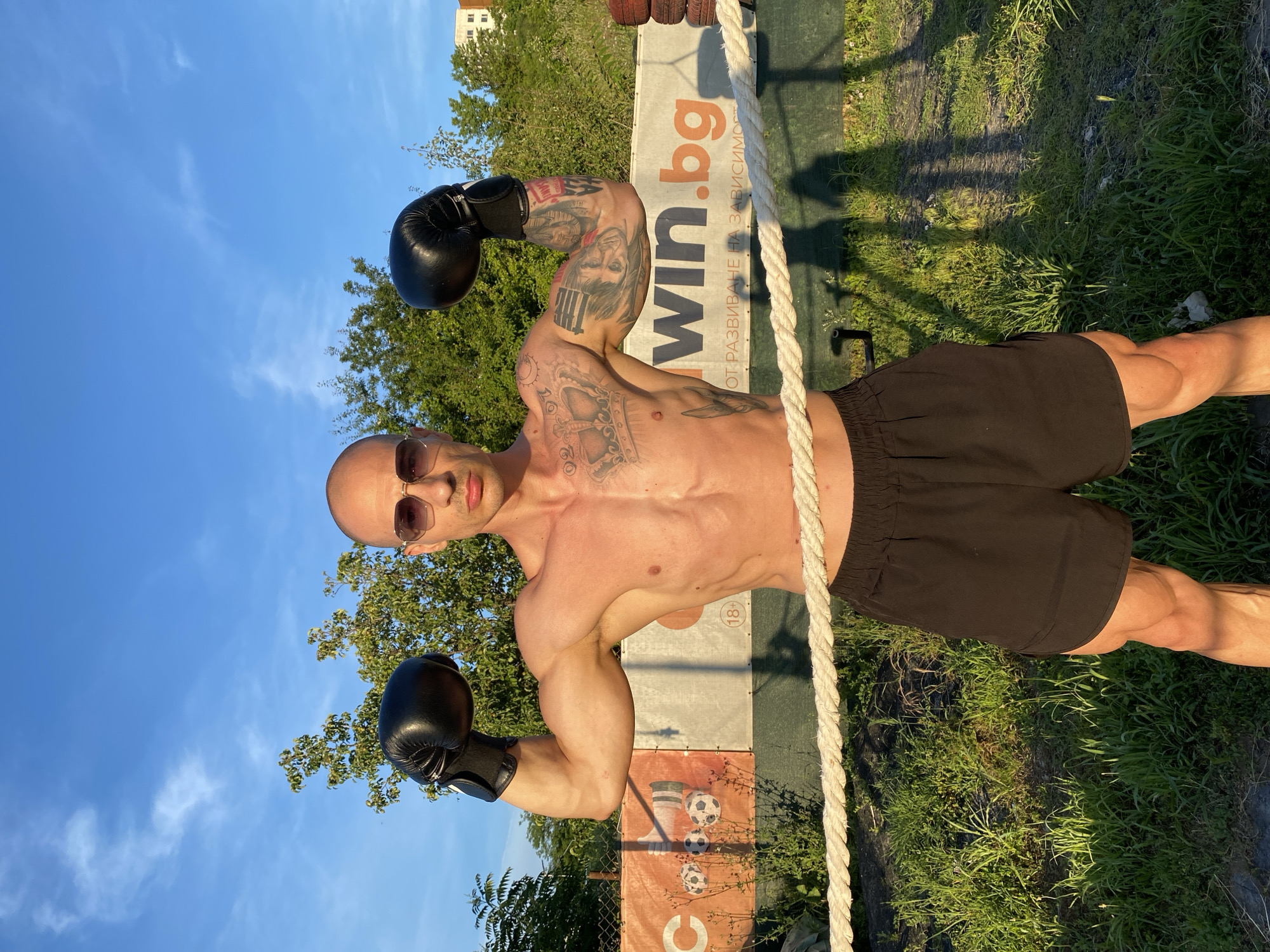 The media file shows a man in a boxing ring outdoors. He is bald, wearing sunglasses, and has black boxing gloves on both hands, which he is flexing. His torso is bare, revealing tattoos on his arms and chest. He is wearing black shorts.
The setting appears to be a boxing ring with ropes and a banner in the background. The banner has text on it, including "win.bg" and some Cyrillic text. There are also graphics of footballs and a boot on the banner. The ground is covered in grass, and trees are visible behind the ring. The sky is blue with some wispy clouds, suggesting it is daytime. The sunlight casts shadows on the grass, indicating it could be late afternoon or early morning.
The man's pose and expression suggest confidence and strength. The overall atmosphere is athletic and energetic, possibly related to a training session or promotional event for boxing. The presence of the "win.bg" text suggests a connection to a Bulgarian website, and the 18+ symbol on the banner indicates a potential age restriction for advertised content. The location context of Pleven, Bulgaria, aligns with the Cyrillic text on the banner.