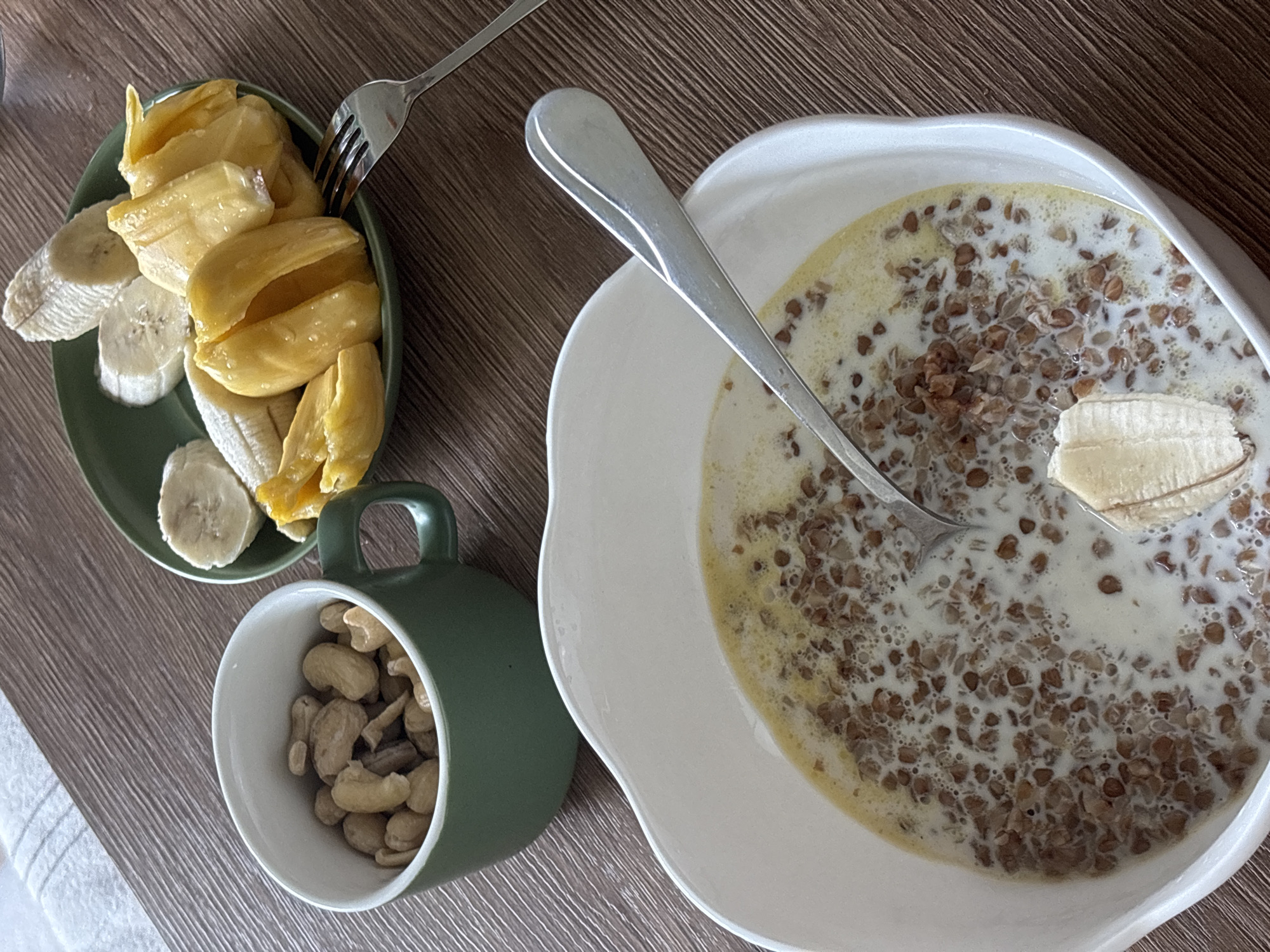 An overhead shot captures a healthy meal or snack spread on a wooden table. In the foreground, a large white bowl contains cooked buckwheat groats submerged in milk, with a silver spoon resting inside and a single banana slice positioned at the edge. To the left, a smaller, dark green plate holds several fresh banana slices and multiple vibrant yellow segments of what appears to be jackfruit. A silver fork is placed on this fruit plate. Below the fruit, a dark green mug is filled to the brim with cashew nuts. A white, textured fabric is partially visible on the far left side of the table. The overall scene suggests an indoor setting, likely for breakfast or a light meal. The presence of jackfruit and bananas is consistent with the tropical context of Choeng Thale, Thailand. There is no visible text or identifiable people in the image.