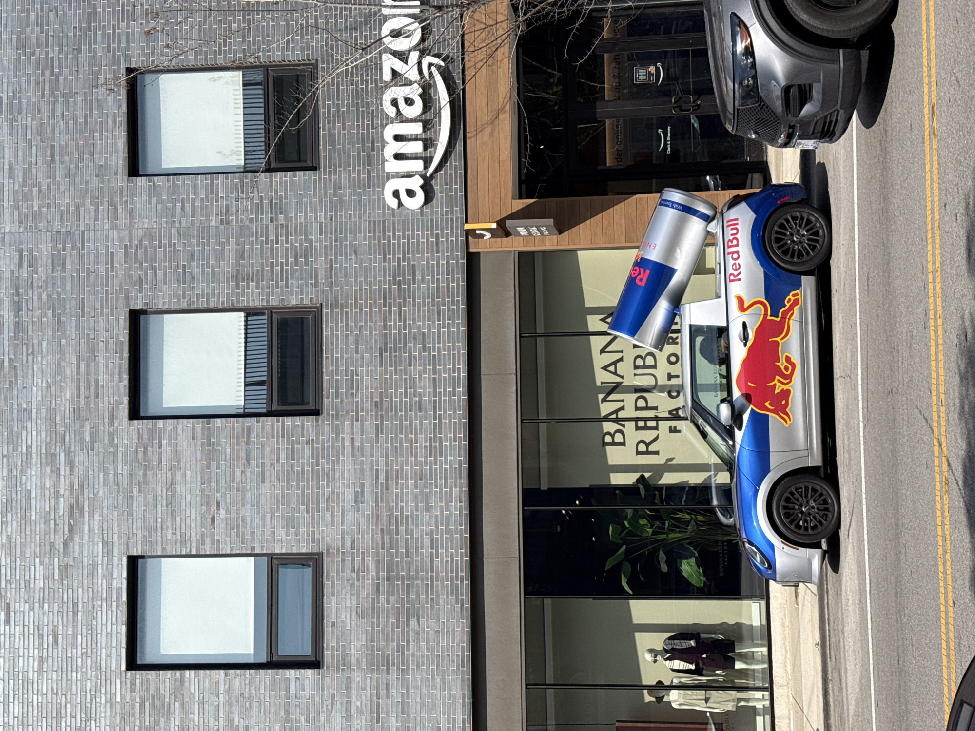 A Mini Cooper convertible is parked on a city street in front of a Banana Republic Factory store and an Amazon store. The car is decorated with a Red Bull logo and a giant Red Bull can is mounted on its roof. The car is silver with blue and red accents. The Red Bull logo features a red bull charging forward. The Mini Cooper has black wheels. In the background, a gray Ford Explorer SUV is parked to the right of the Mini Cooper. The Banana Republic store has large windows displaying mannequins dressed in casual attire. The Amazon store has a wooden facade and glass doors. The stores are located on a street with yellow lane markings. The building behind the stores is made of gray brick. The sky is clear and the sun is shining. The image appears to be taken in Chicago, United States of America.