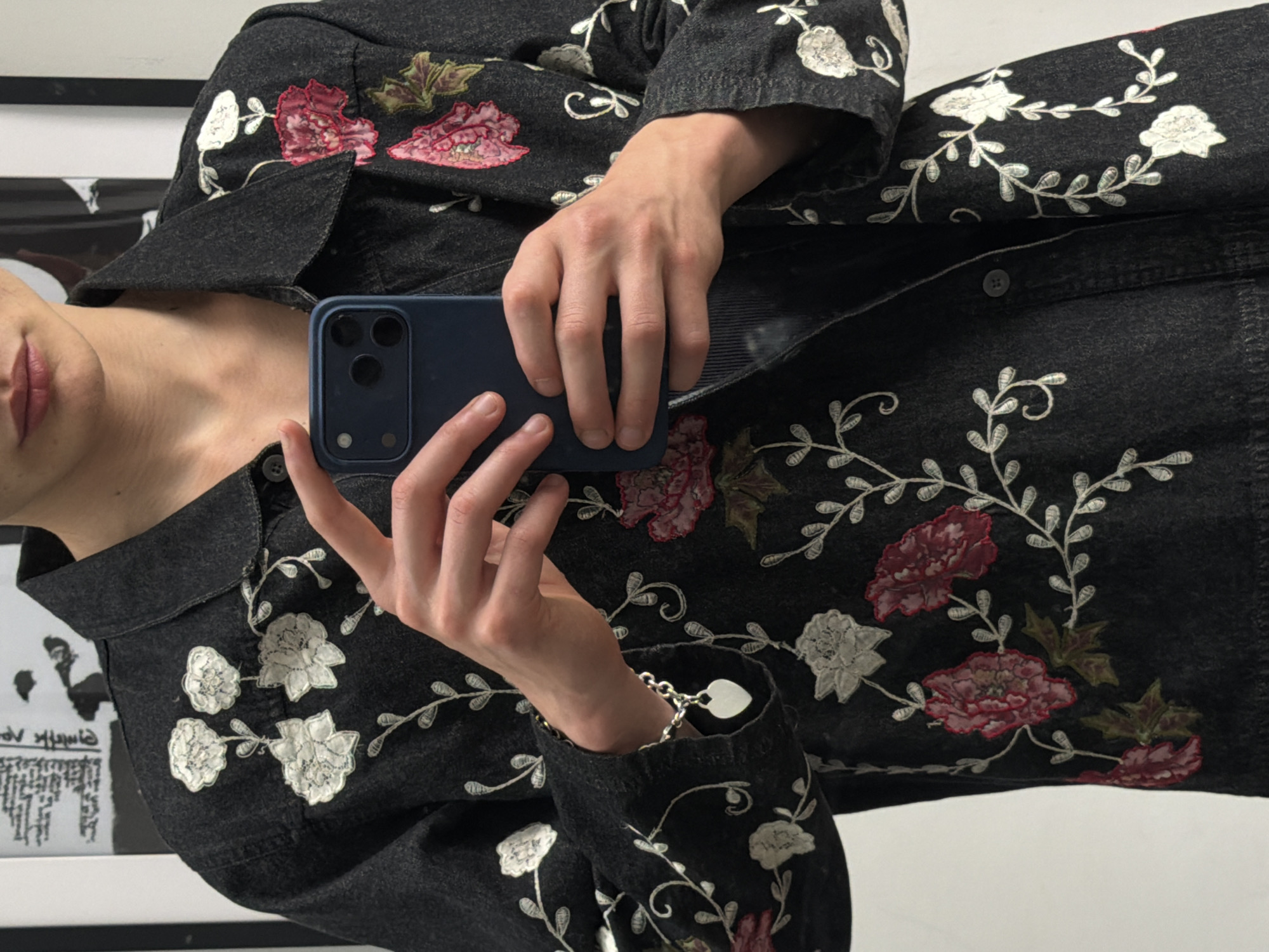 The upper torso of an individual is depicted holding a dark blue smartphone horizontally with both hands, obscuring the face, against a light background. The person wears a dark charcoal or faded black denim-style jacket, heavily embroidered with white and pink-red floral patterns and delicate white vine motifs. A silver-colored chain-link bracelet with a polished, flat silver heart charm adorns the left wrist. The jacket is unbuttoned at the front, revealing a dark, possibly ribbed garment underneath, with a single dark button visible on the left side of the jacket opening. The background consists of a light-colored wall with indistinct, blurry framed items or posters, one showing dark text or abstract shapes on a light ground, and another with a prominent black vertical stripe. There are no visual elements within the image that allow for the identification of a specific geographic location, such as the city of London, United Kingdom.
