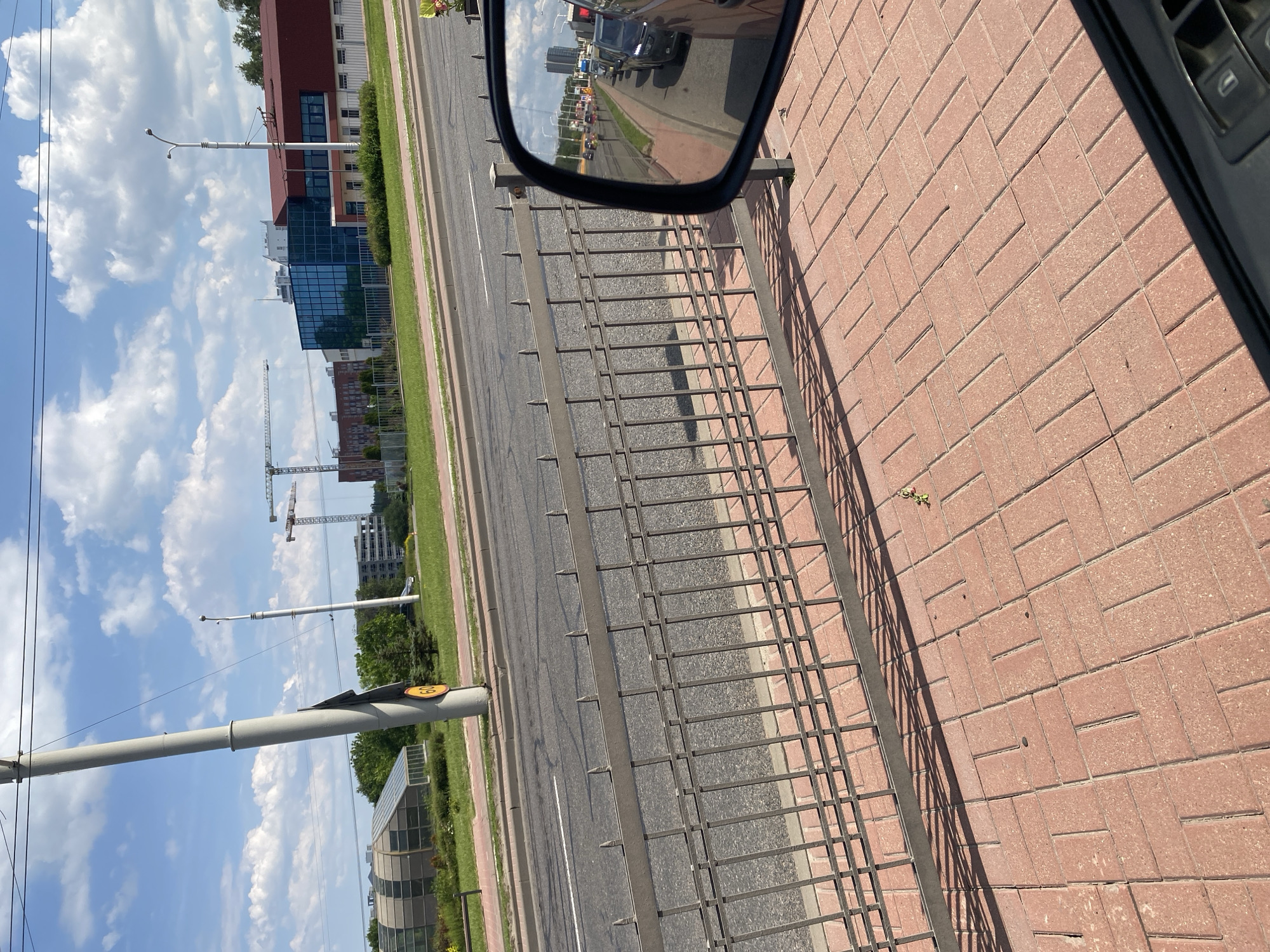 The image shows a view from a car of a street scene. In the foreground, there is a red brick sidewalk and a metal fence. Beyond the fence is a road with several cars driving on it, visible in the wing mirror of the car from which the photo was taken. In the background, there are buildings, trees, and construction cranes. The sky is blue with white clouds. There are no people clearly visible in the image. The scene appears to be in a city.