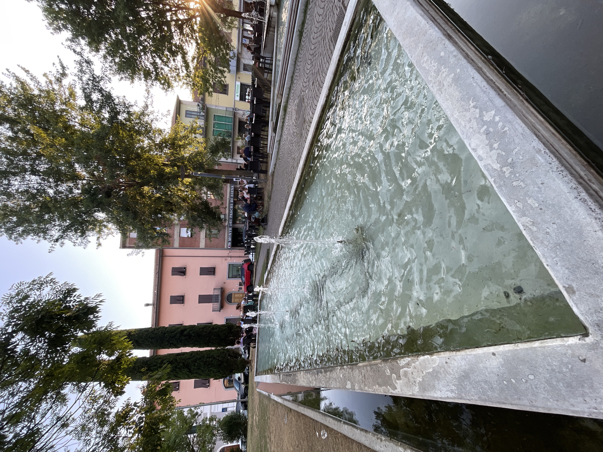 The image depicts a public fountain with water jets in the foreground, set against a backdrop of buildings and trees. People are seated at tables on a paved area behind the fountain, suggesting a public square or park area. Several trees are visible, some tall and slender cypress trees and others with fuller canopies. The buildings are a mix of pastel colors, with balconies and windows. Sunlight is visible, casting a warm glow and lens flare in the upper right corner. The fountain is long and rectangular, with concrete edges and clear water. The overall scene suggests a relaxed outdoor setting in a European town. Based on visual context, the location appears to be Piazza Rabadan in Altopascio, Italy, a known area with fountains and outdoor seating for cafes and restaurants.