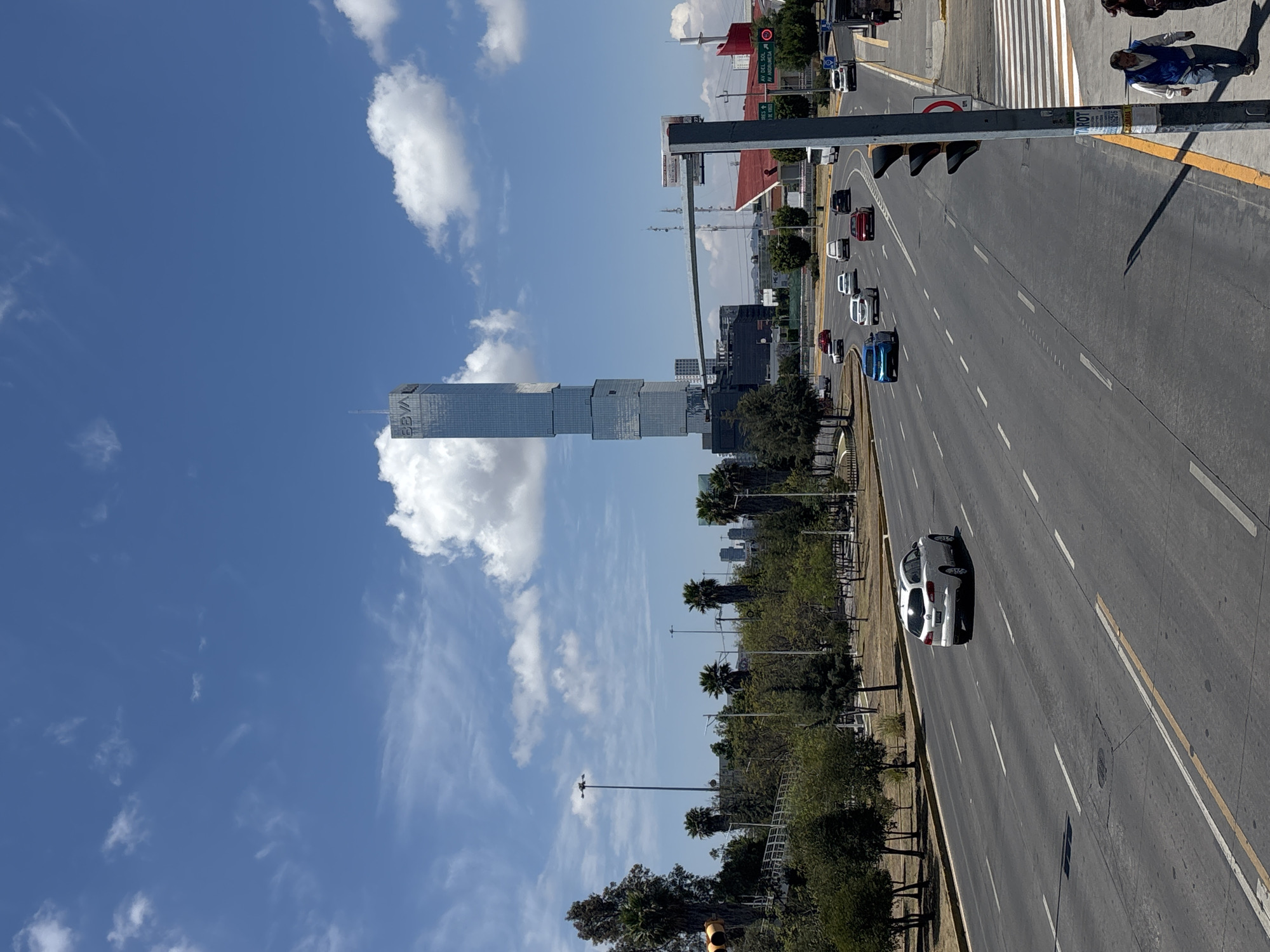 The image depicts a view of the BBVA Tower in San Bernardino Tlaxcalancingo, Mexico. The tower is visible in the background, rising high into a blue sky with scattered clouds. A multi-lane road stretches into the distance with vehicles traveling along it. Some pedestrians are visible on a sidewalk near a crosswalk. Vegetation is located along the roadside, including trees and greenery.