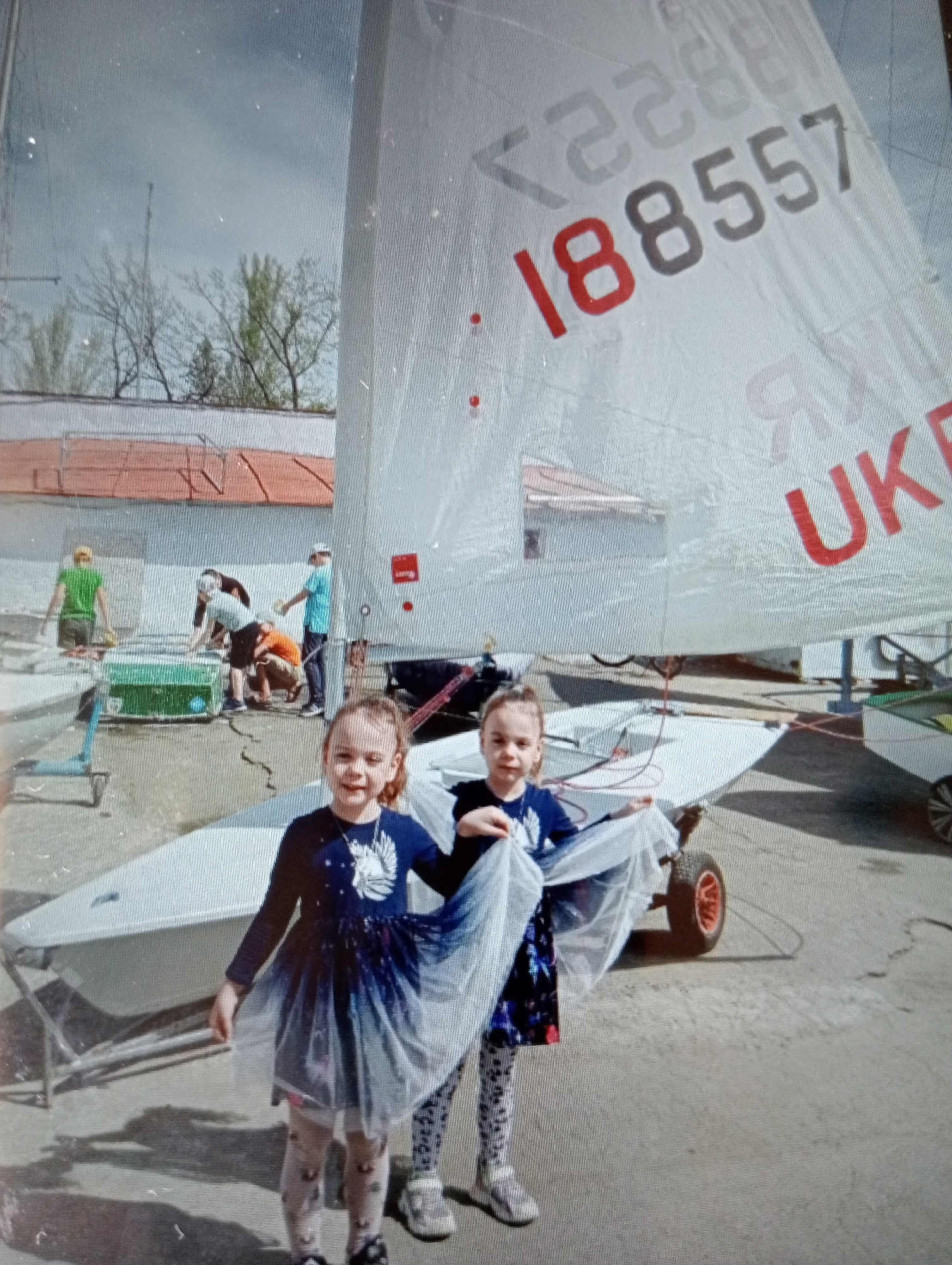 The image depicts a sunny outdoor scene, likely a marina or boatyard, with a large sailboat as the focal point. Two young girls, dressed in festive outfits, are positioned in the foreground, holding up the sail of a smaller boat. They are smiling at the camera. In the background, several adults appear to be working on or around boats, with a green container visible near them. The environment features a paved surface, a building with a red roof, and trees in the distance under a cloudy sky. The sail of the large sailboat prominently displays the numbers "188557" and the letters "UKR." The overall atmosphere suggests a cheerful event, possibly a sailing competition or festival.
