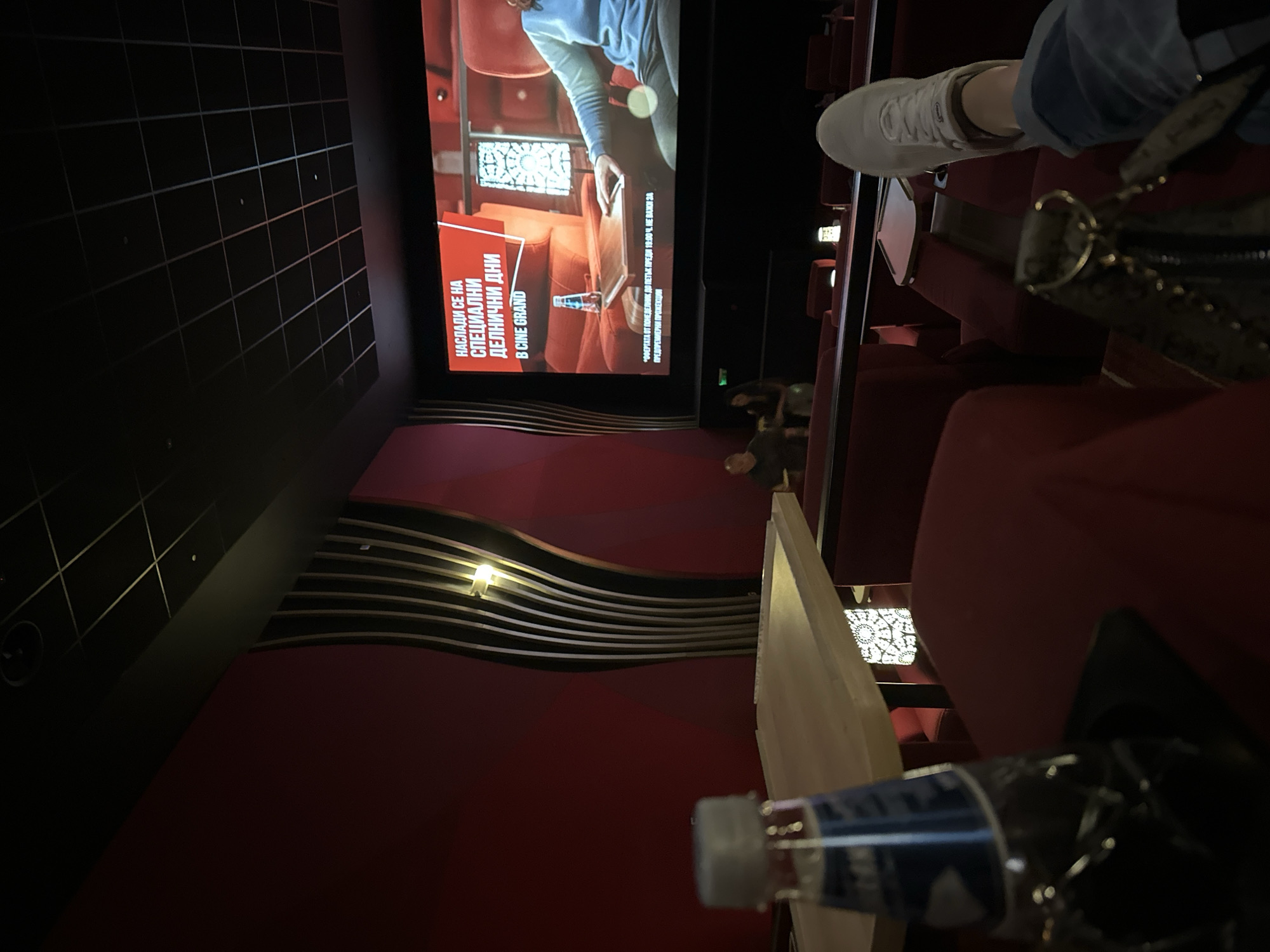 This image depicts a dimly lit, upscale cinema hall at Cine Grand, likely within the Ring Mall location in Sofia, Bulgaria. The perspective is from a seated individual looking towards the large projection screen.
In the foreground, a plastic water bottle with a blue label and white cap rests on a light-colored wooden table to the left. To the right, a person's left foot, adorned with a white sneaker and blue jeans, is visible, suggesting a relaxed posture with the foot possibly resting on an adjacent seat or footrest. A dark-colored handbag with gold-toned hardware is partially visible in the bottom right corner.
The midground features rows of plush, deep red cinema recliners, each equipped with an individual wooden side table. Two other patrons are vaguely visible in the lower rows to the left of the screen, appearing as dark silhouettes.
The prominent cinema screen displays an advertisement in Bulgarian. The text in a red banner reads: "НАСЛАДИ СЕ НА СПЕЦИАЛНИ ДЕЛНИЧНИ ДНИ В CINE GRAND" (ENJOY SPECIAL WEEKDAY DAYS AT CINE GRAND). Smaller text below details the terms: "*ОФЕРТАТА ОТ ПОНЕДЕЛНИК ДО ПЕТЪК ПРЕДИ 18:00 Ч. НЕ ВАЖИ ЗА ПРЕДПРЕМИЕРНИ ПРОЖЕКЦИИ" (*THE OFFER FROM MONDAY TO FRIDAY BEFORE 6:00 PM IS NOT VALID FOR PRE-PREMIERE SCREENINGS). The visual content of the advertisement shows a luxurious interior scene with red armchairs, a wooden table, a decorative lamp, and a person in a light blue top with a similar water bottle.
The environment of the hall is characterized by deep red walls, possibly upholstered, and tall, curved decorative panels with alternating dark and light vertical strips, visible on both sides of the screen. A warm, yellow light fixture is integrated into the right-side paneling. The ceiling is dark and features a visible grid pattern, with small, recessed lights distributed across it. The overall ambiance is designed for a premium viewing experience.