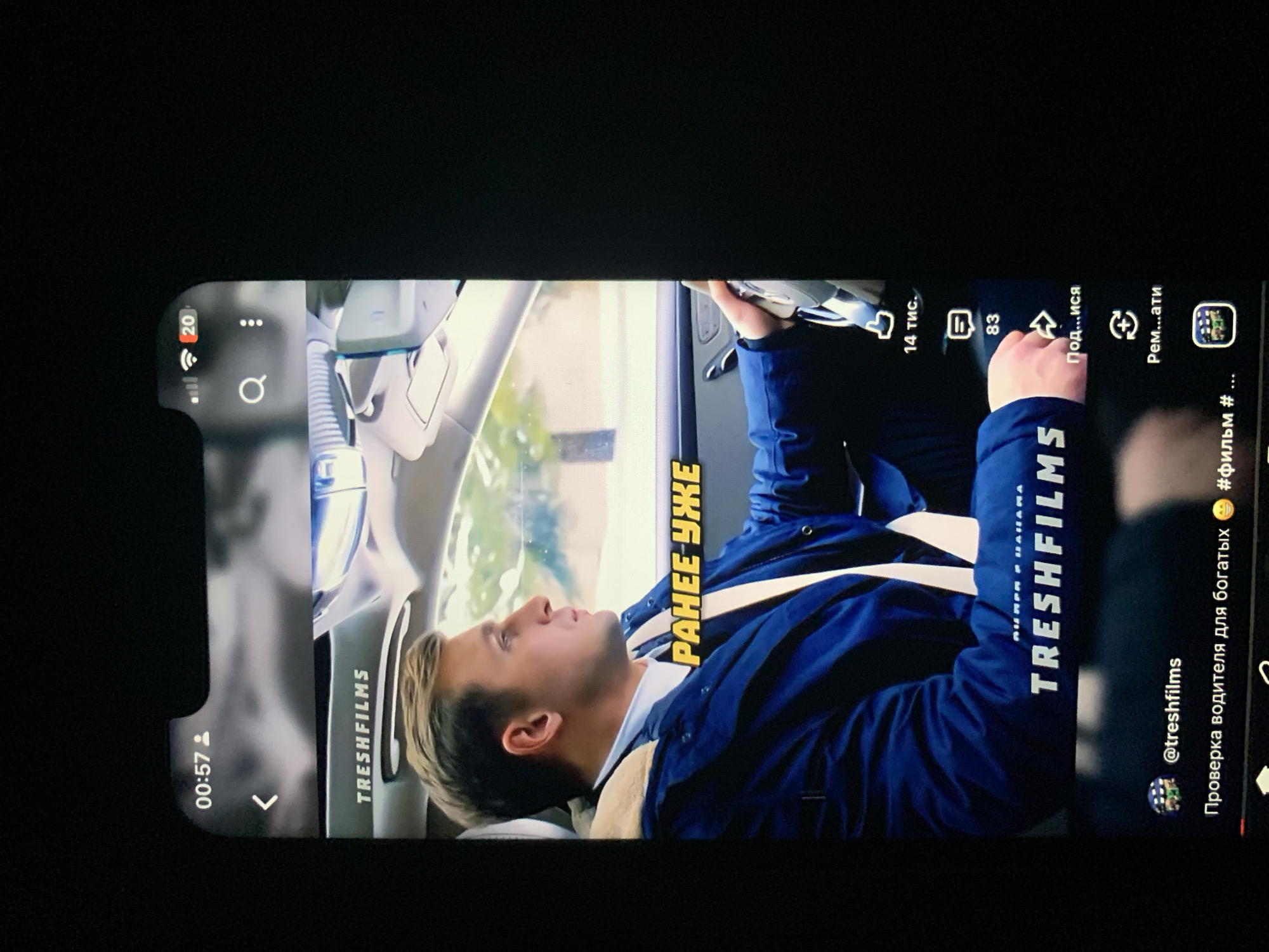The image shows a close-up of a smartphone screen displaying a video. The video features a man in a blue jacket with a fur-lined collar, seated in the driver's seat of a car. He is looking to his right, with his hands on the steering wheel. Yellow text overlays the video, reading "РАНЕЕ УЖЕ". Below the video, there are social media interface elements, including a username "@treshtfilms", a caption in Russian that translates to "Driver check for the rich", and hashtags. The overall scene is viewed from a slightly elevated angle, suggesting the phone is being held or placed in a way that captures this perspective. There is no visible indication of the city of Zhmerynka, Ukraine, or any specific geographic location within the image itself.