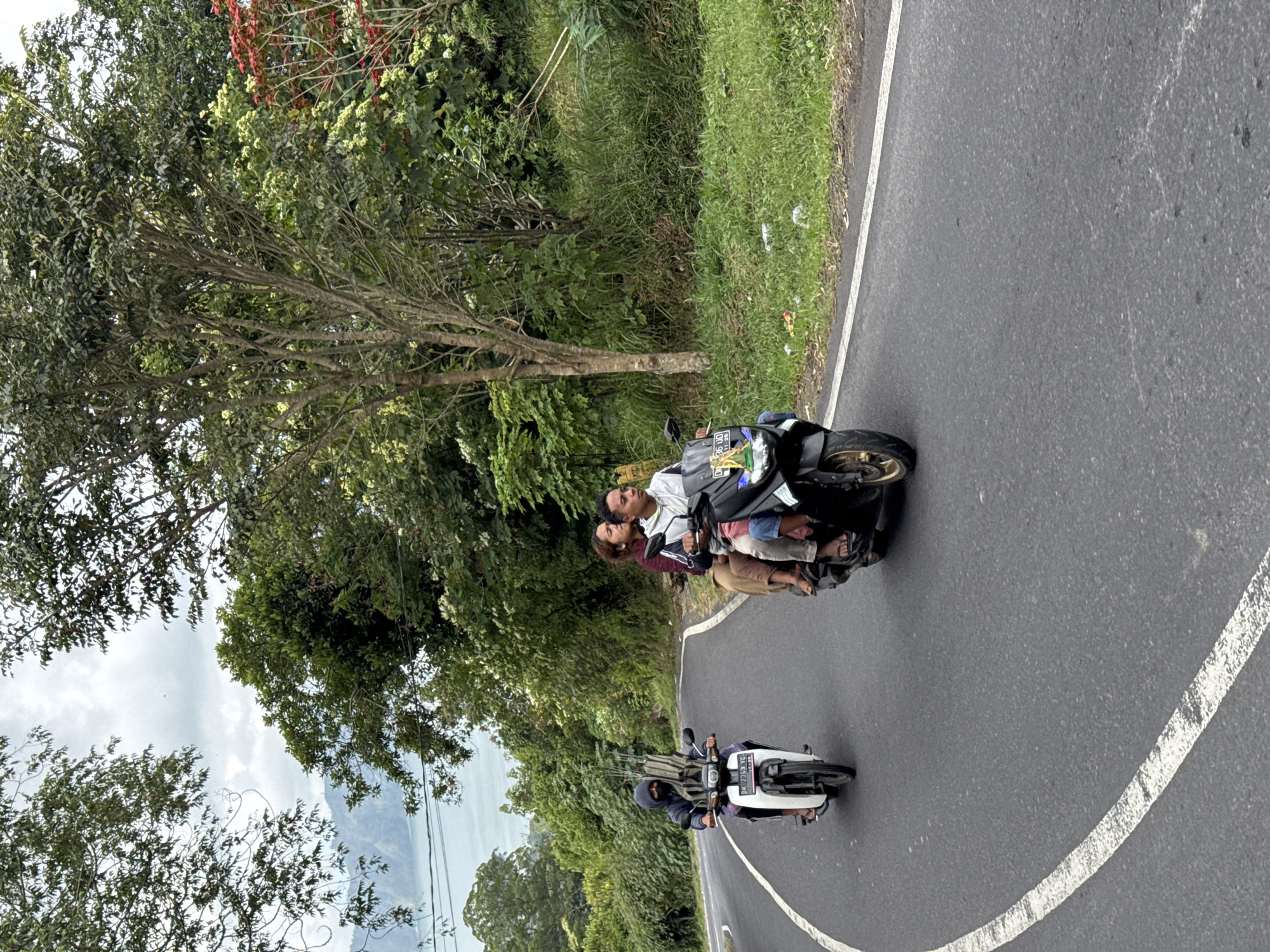 This media file depicts a scene on a winding road in Kintamani, Indonesia. Two motorcycles are visible, one in the foreground and another further up the road. The motorcycle in the foreground is white with black accents, and a person is riding it, wearing a dark hooded jacket and gloves. The motorcycle further up the road is black and has two people on it. The person in front is wearing a white shirt and the person behind them is wearing a maroon shirt. They appear to be looking at something to their left.
The setting is lush and green, with dense vegetation and trees lining the road. The road itself is paved with a white line marking the edge. In the background, a mountainous landscape and a body of water can be seen, suggesting a scenic, elevated location. The sky appears overcast, indicating a possibility of rain or a cloudy day. The time of day is not clearly discernible but appears to be daytime. The overall mood seems relaxed and perhaps adventurous, given the mode of transport and the scenic route.
Visible text includes license plates on both motorcycles. The front motorcycle has a license plate with the text "DN 2853 VC". The license plate on the black motorcycle is partially obscured but appears to have "GN 998D" visible.