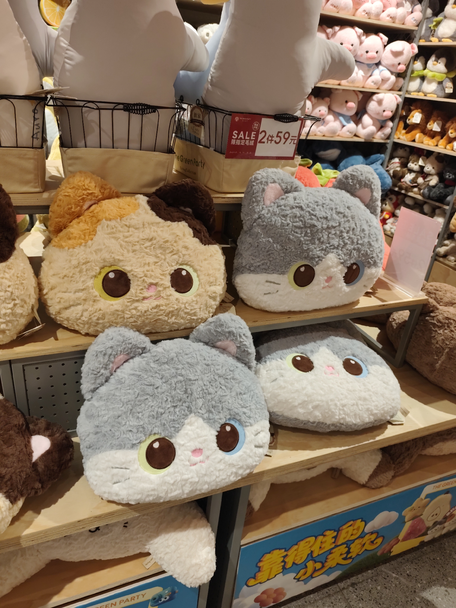 This is a close-up, eye-level, indoor photograph of a retail display of stuffed animals in Daqing, China. The foreground features several plush cat-shaped pillows. On the top shelf, from left to right, are a calico cat pillow and a gray cat pillow. On the shelf below, from left to right, are another gray cat pillow and a white and gray cat pillow. Behind these, a large white plush animal, possibly a shark or whale, is visible. To the right, shelves filled with various other plush toys, including pink pigs and stuffed lions, are visible. A price tag with Chinese characters and the word "SALE" is attached to a black wire basket on the top shelf. The overall environment appears to be a toy store or department store section.