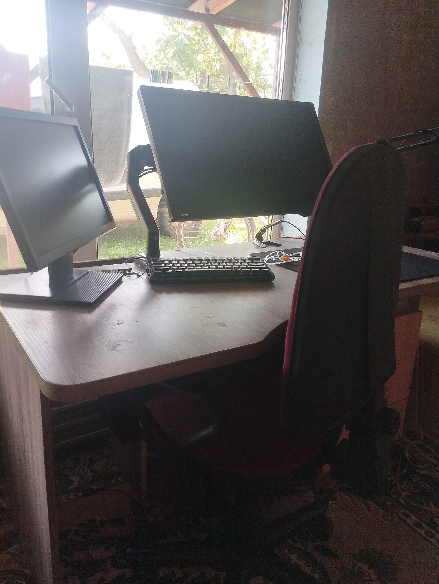 The image depicts a desk with two computer monitors, a keyboard, and a chair in what appears to be a residential setting in Ustyvytsia, Ukraine. The desk is positioned near a window with natural light illuminating the scene. The monitors are mounted on adjustable arms, suggesting a workspace. The keyboard is black and has a standard layout. The chair is dark red and appears to be an office-style chair. The background shows a glimpse of the outdoors through the window, with trees and sunlight suggesting a daytime setting. The overall environment appears to be a room with a carpeted floor. There are no people visible in the image.